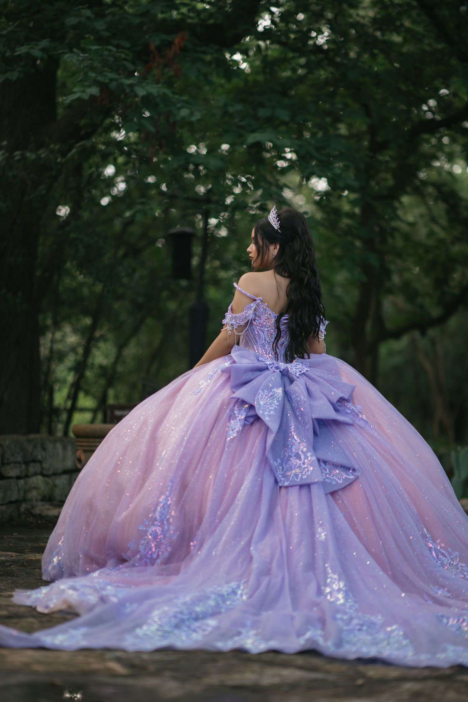 A stunning purple and pink quinceañera dress with a large bow detail shown from behind in a garden.