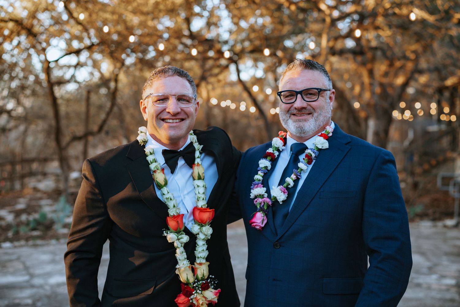 Wedding party members wearing floral garlands and formal attire outdoors at sunset.