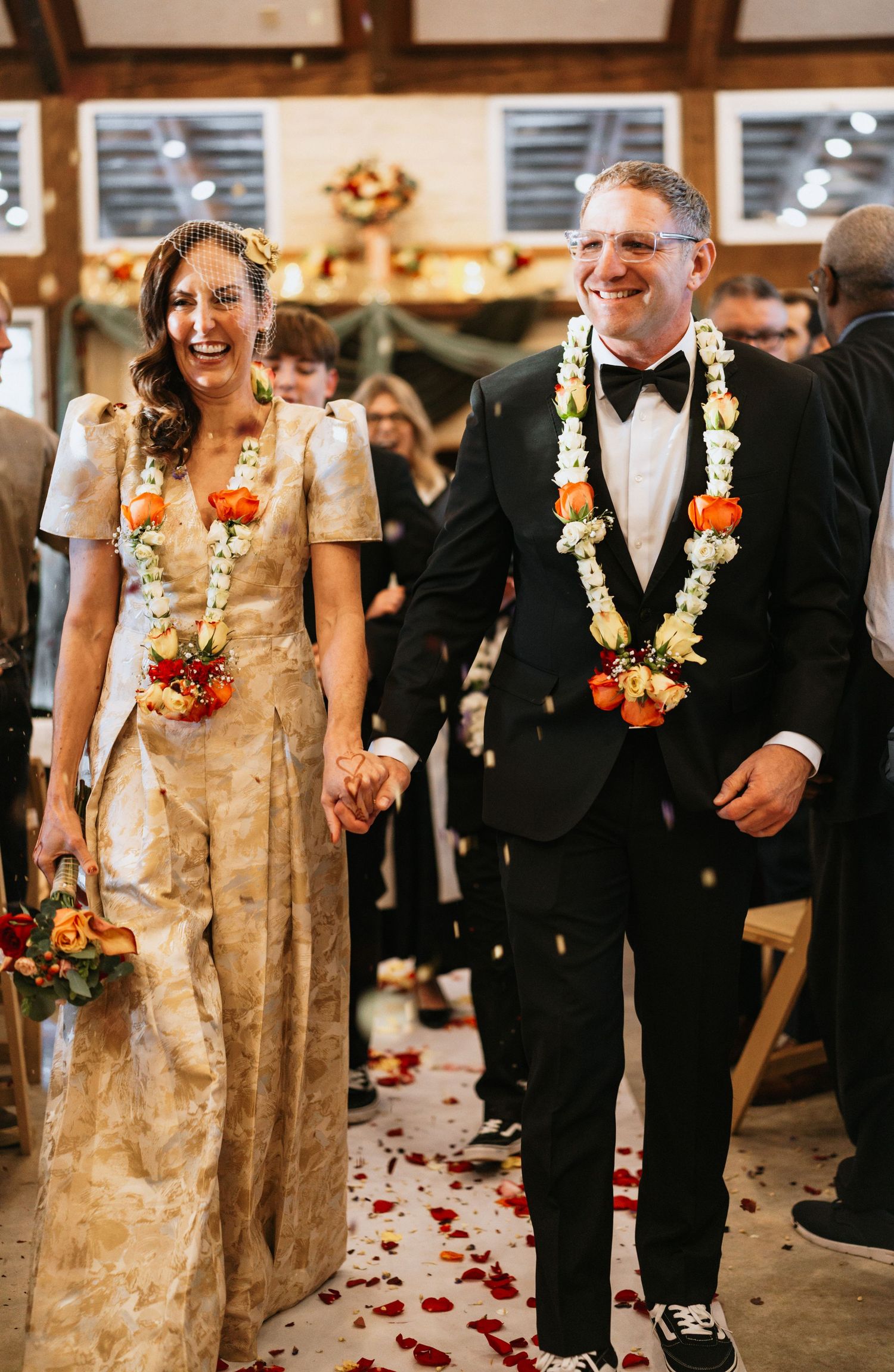 Joyful couple wearing floral leis walks down aisle strewn with rose petals.