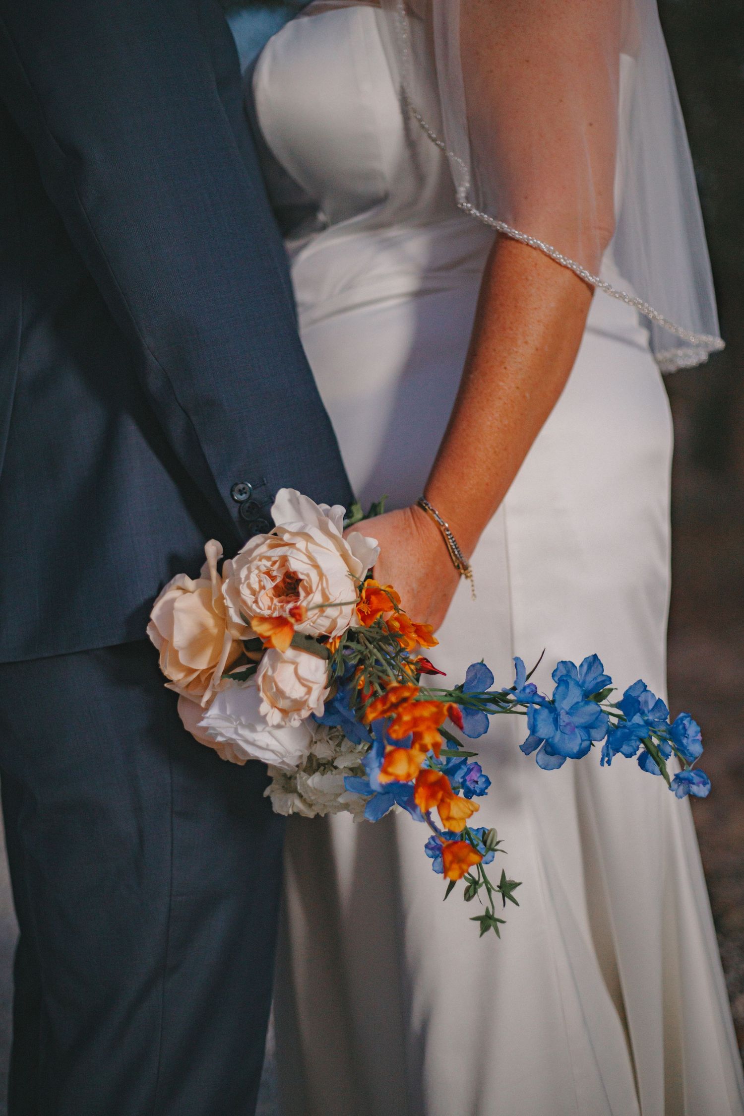 Close-up of wedding bouquet featuring blue, orange and white flowers against white dress.