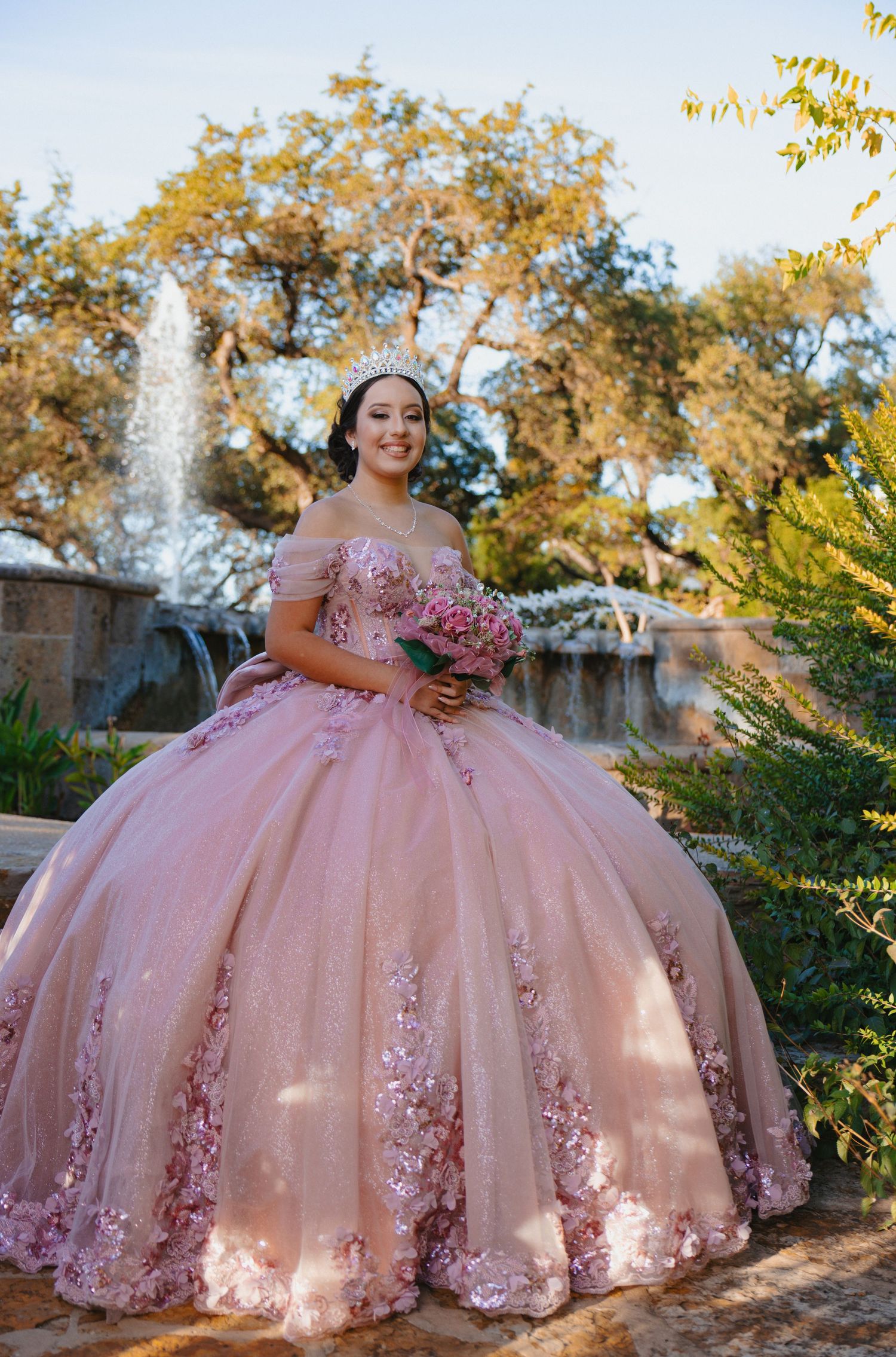 Stunning pink quinceañera ball gown with floral appliques photographed in an outdoor garden setting at sunset.