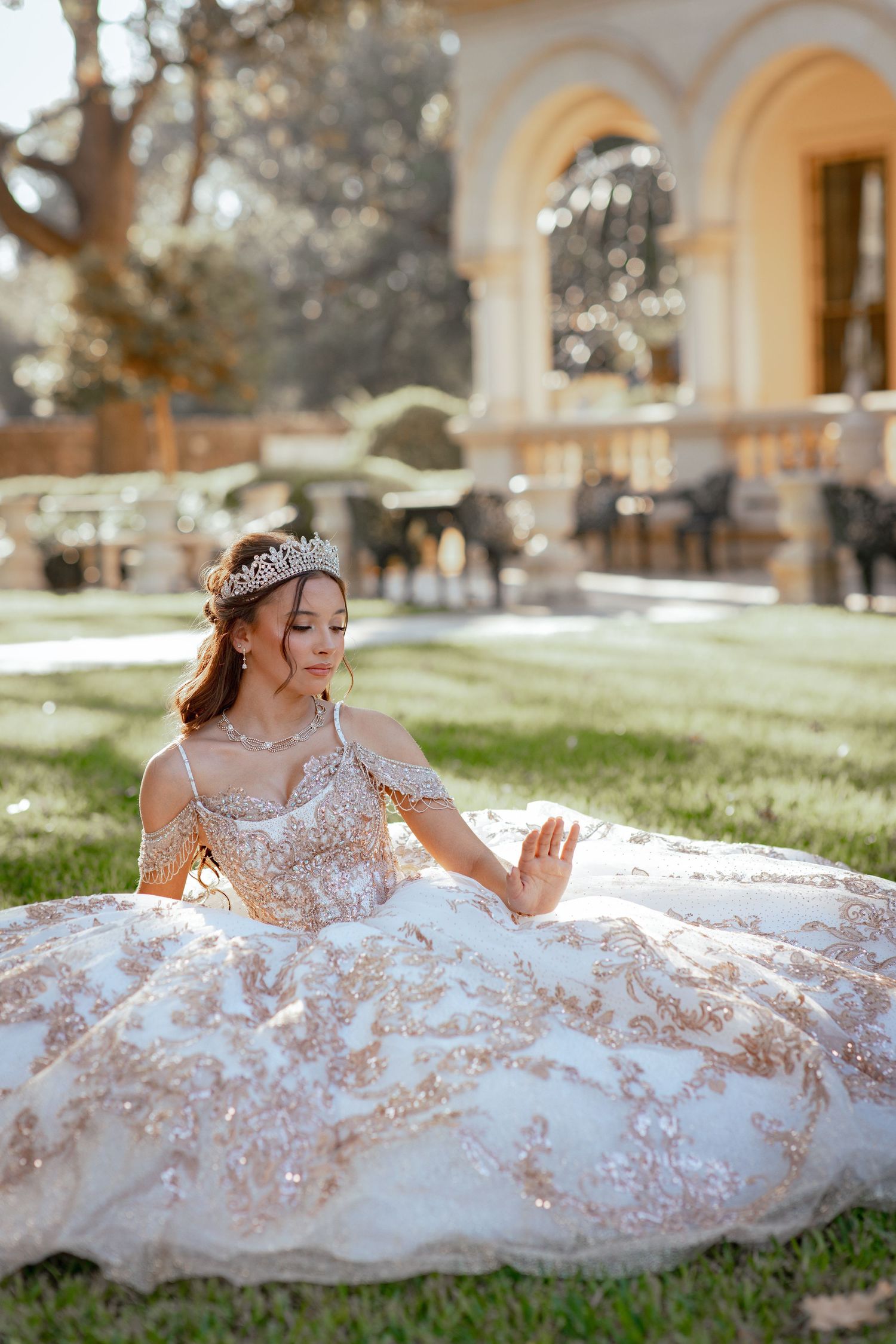 Elegant white and rose gold ball gown with intricate floral details sprawled on grass near ornate archways.