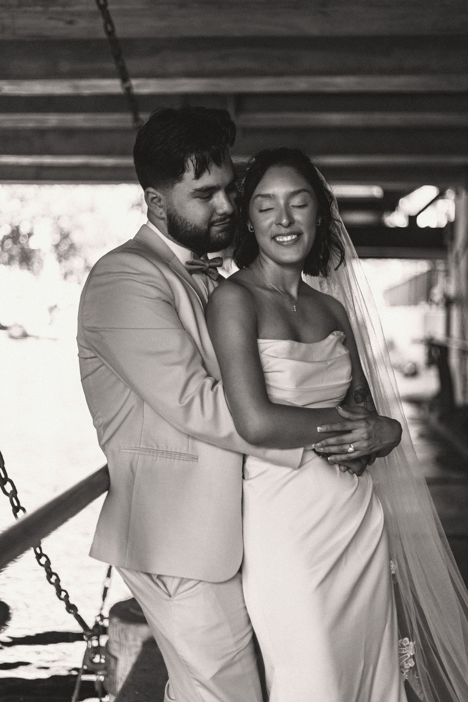 A happy newlywed couple embracing in black and white, with bride in white dress and groom in light colored suit.