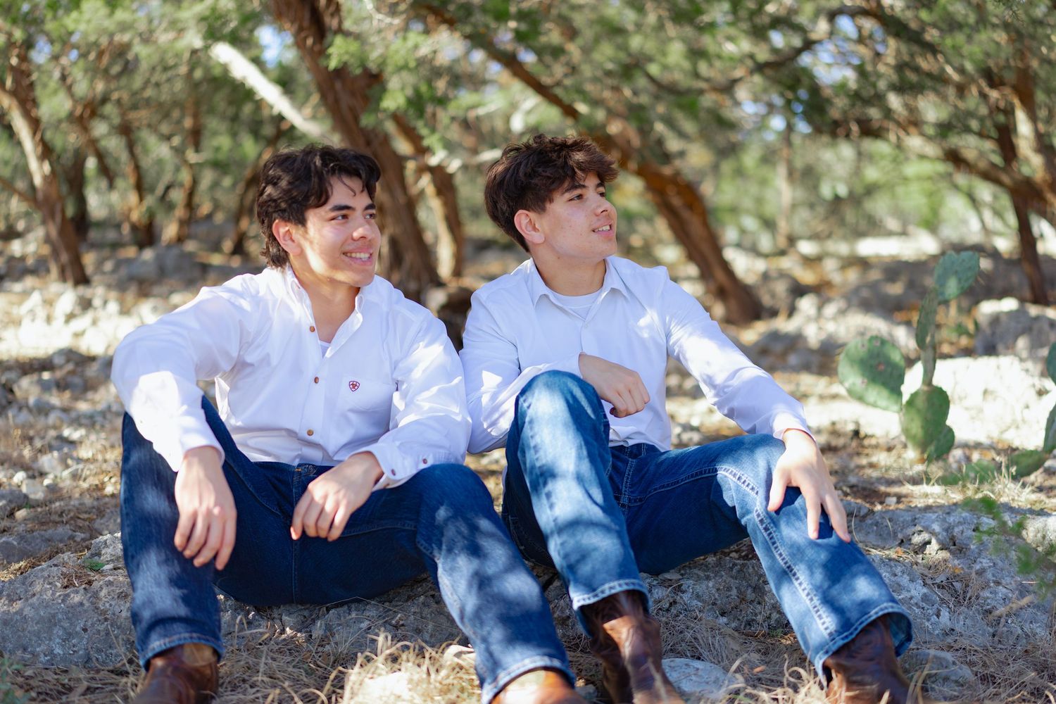 Two young men in matching white shirts and jeans relaxing outdoors among trees.
