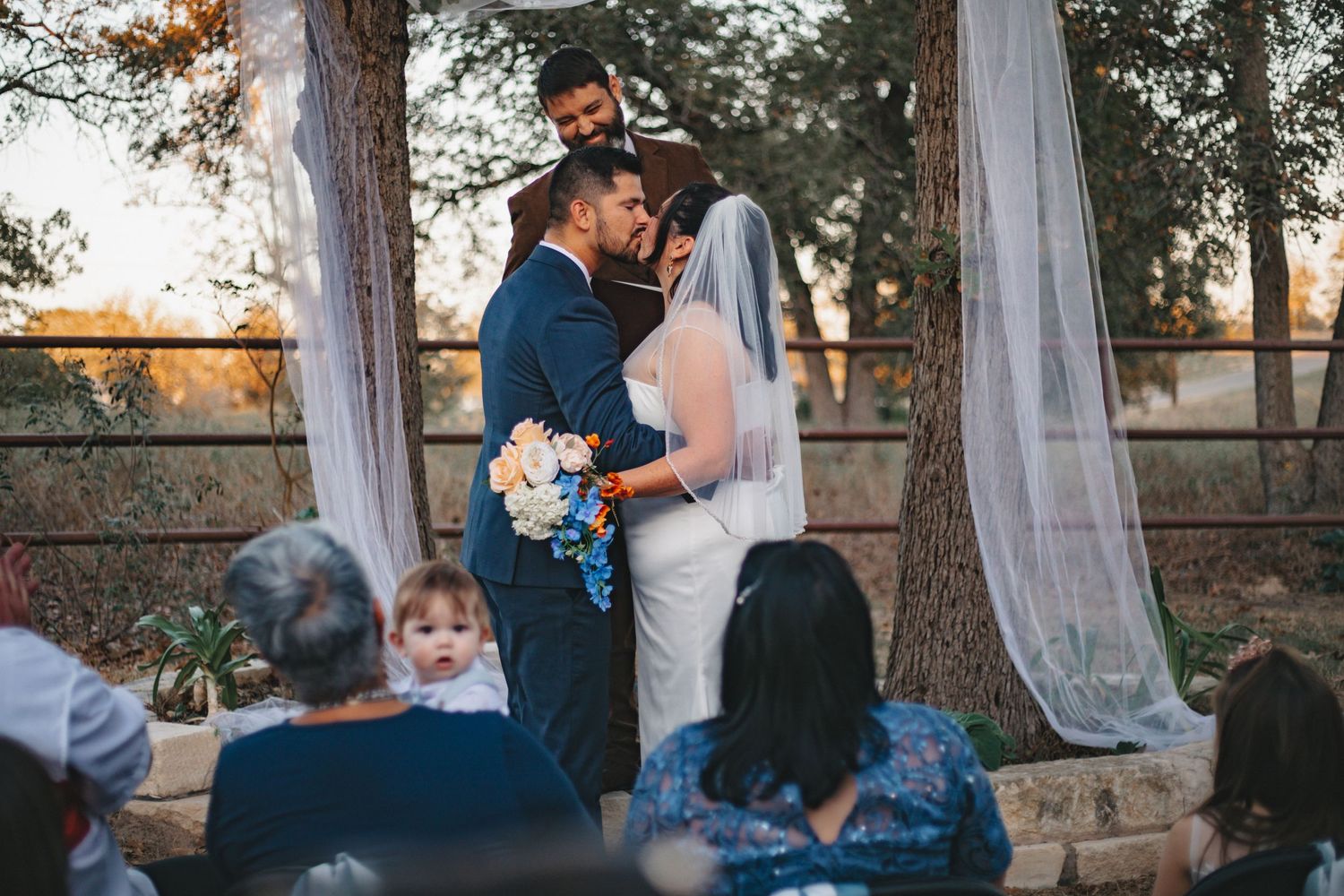 Wedding couple and flower girl share a tender moment under draped fabric and trees at outdoor ceremony.