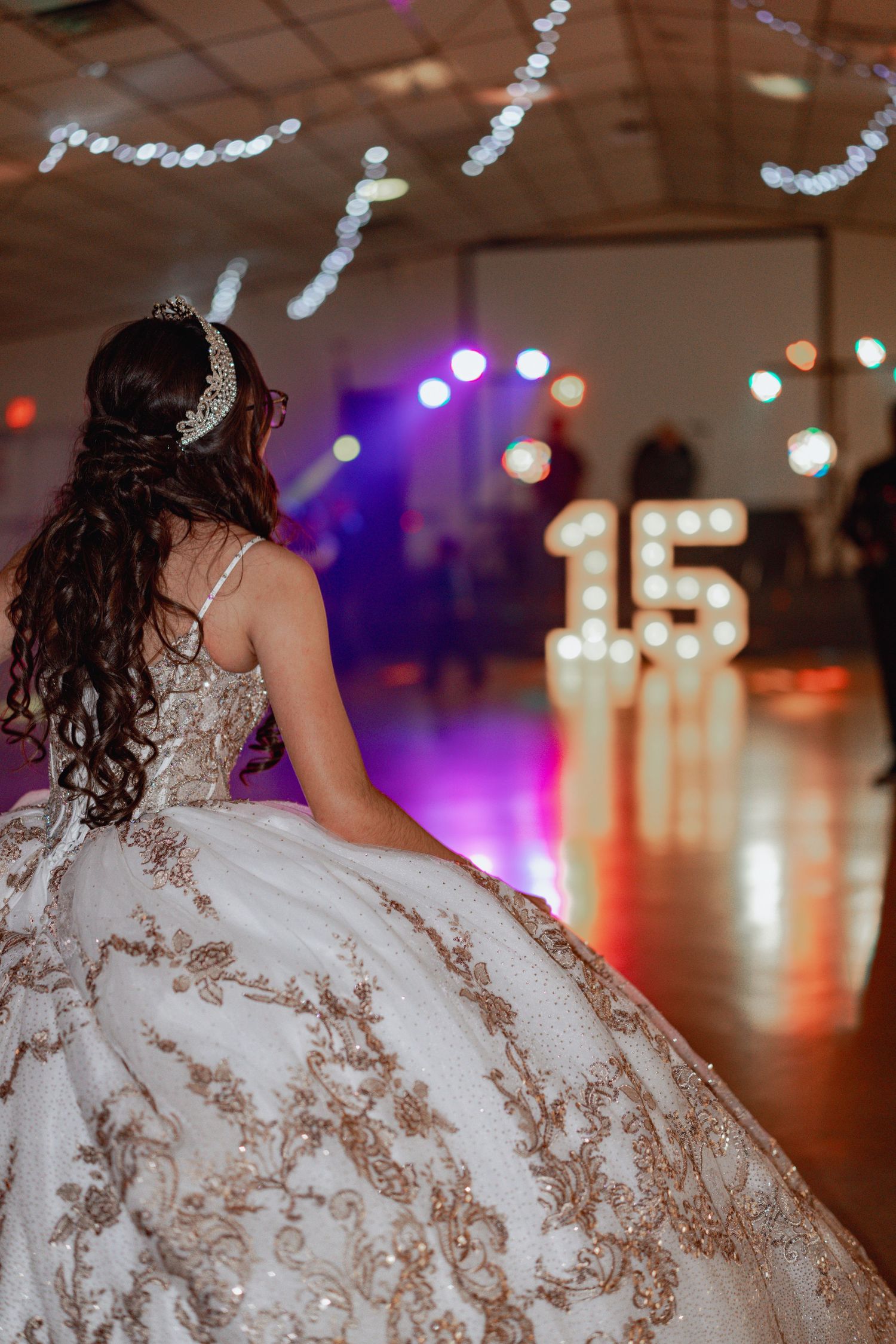 A person in an elaborate white ball gown with beaded details twirls on a dance floor under party lights.