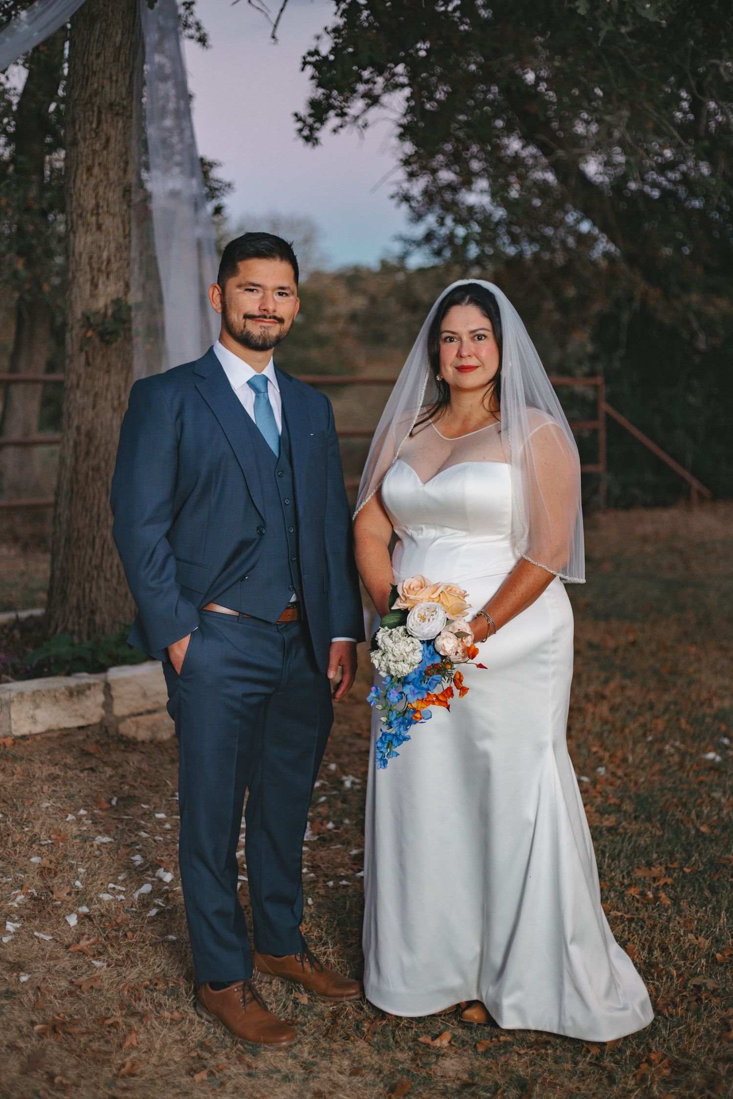 Couple poses for wedding portraits in outdoor setting with trees and white draping.