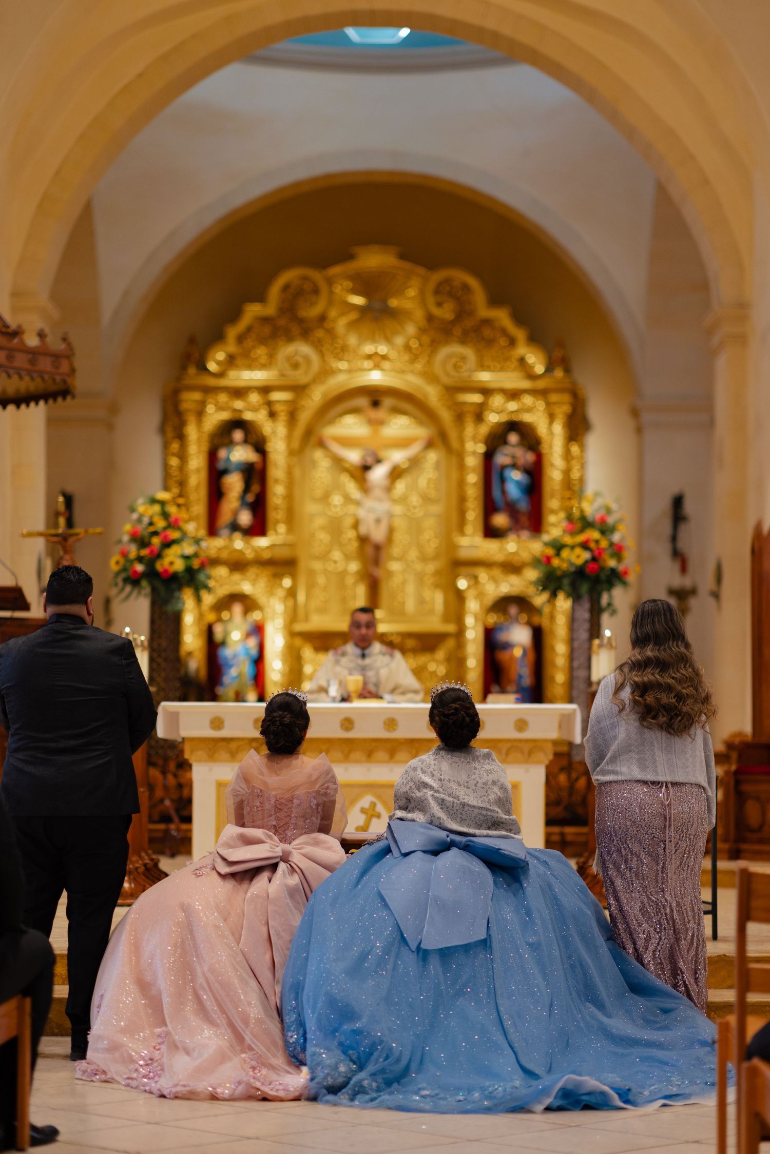 Two people in elegant ball gowns with large bows attend a religious ceremony in an ornate Catholic church.