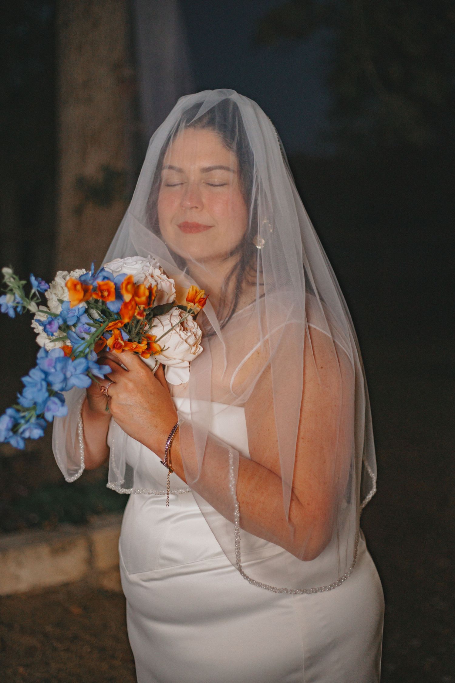 Bride in white dress smells vibrant bouquet with orange and blue flowers against dark background.