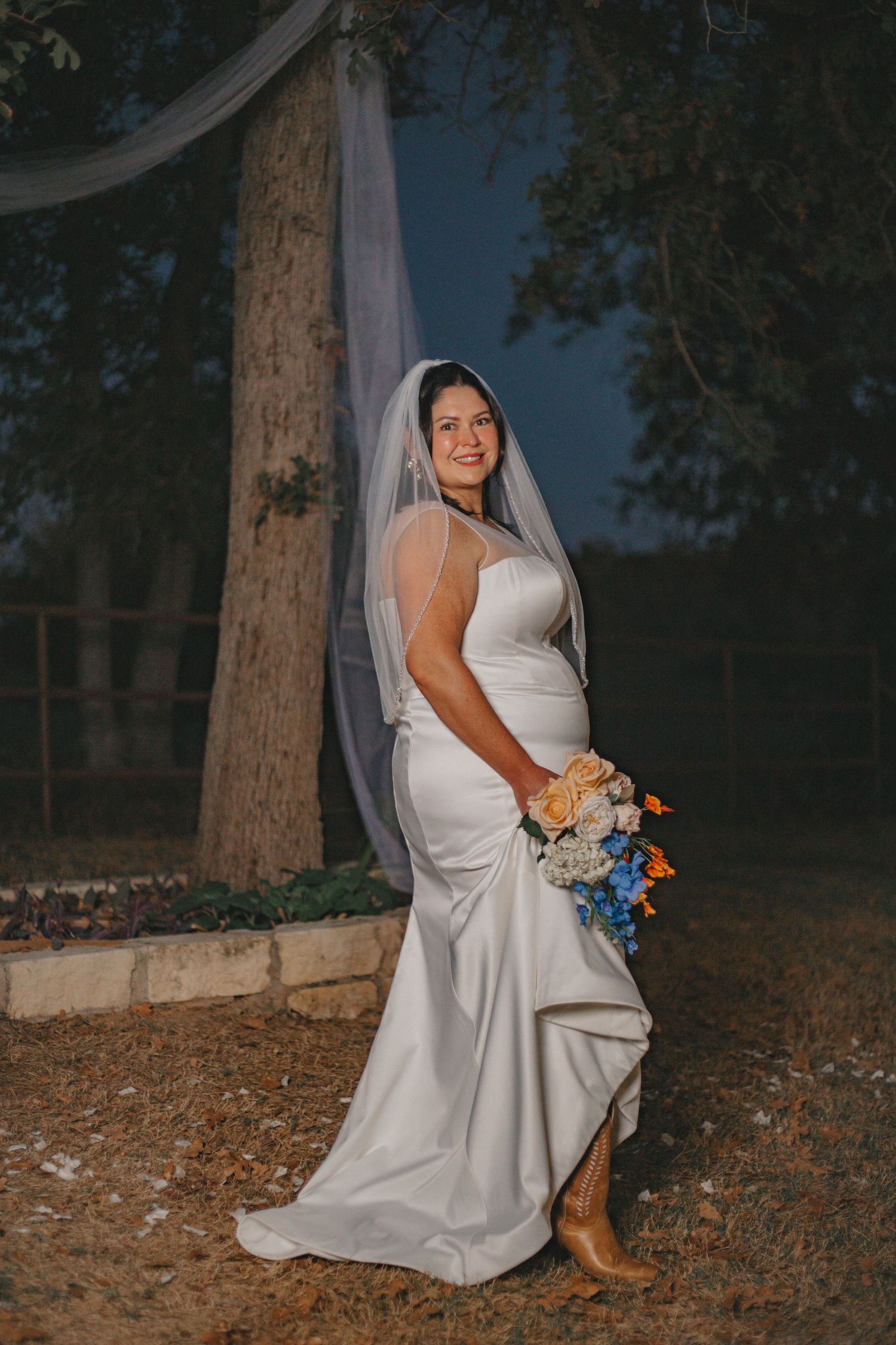 Bride in flowing white dress poses outdoors among trees at dusk holding colorful bouquet.