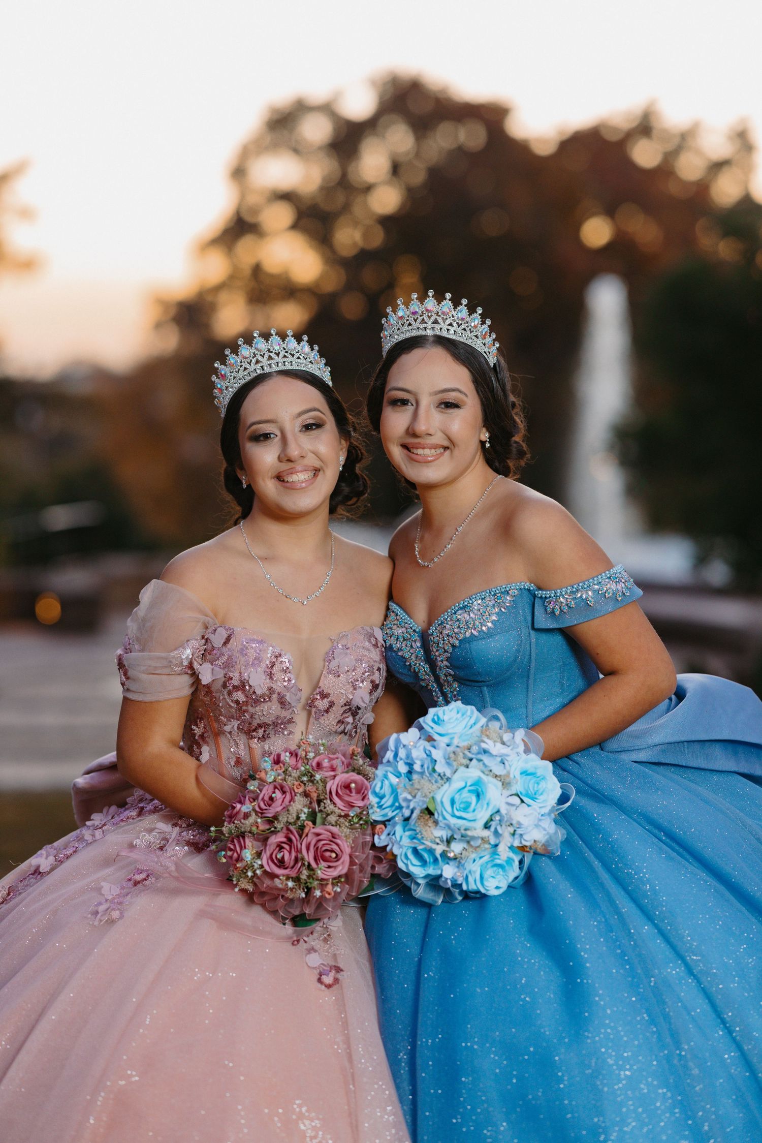 Two formal ball gowns in pink and blue with matching tiaras and bouquets at sunset.