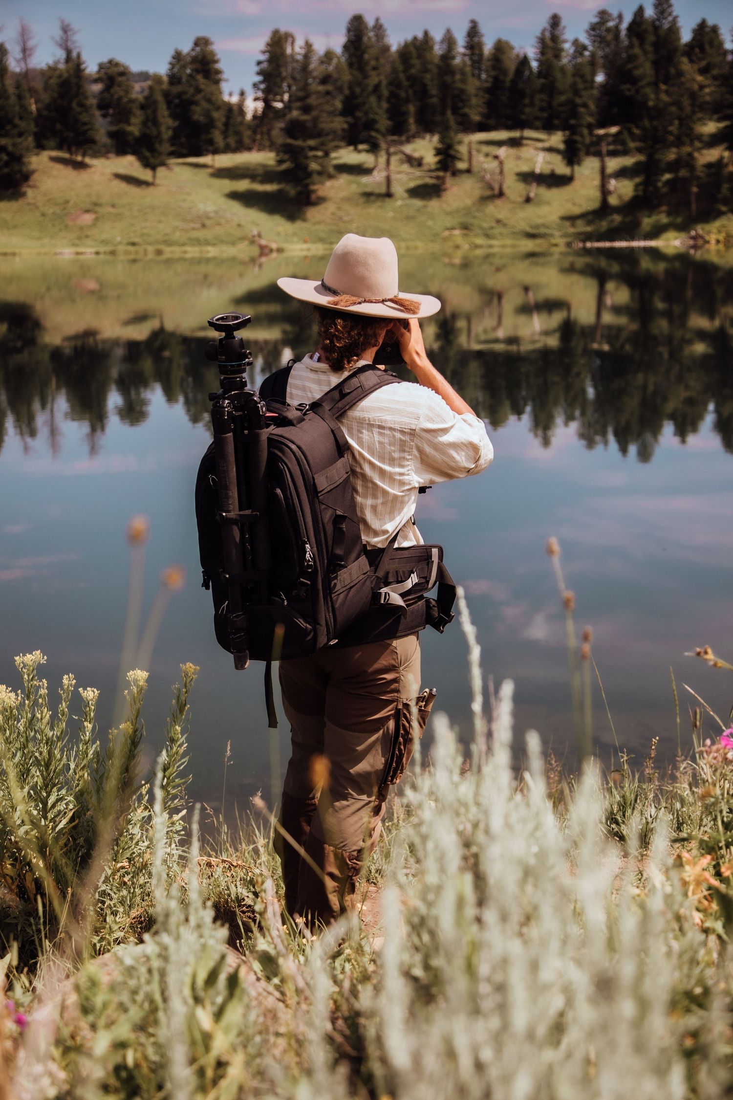 Female photographer with backpack and hat stands by reflective mountain lake surrounded by pine trees.