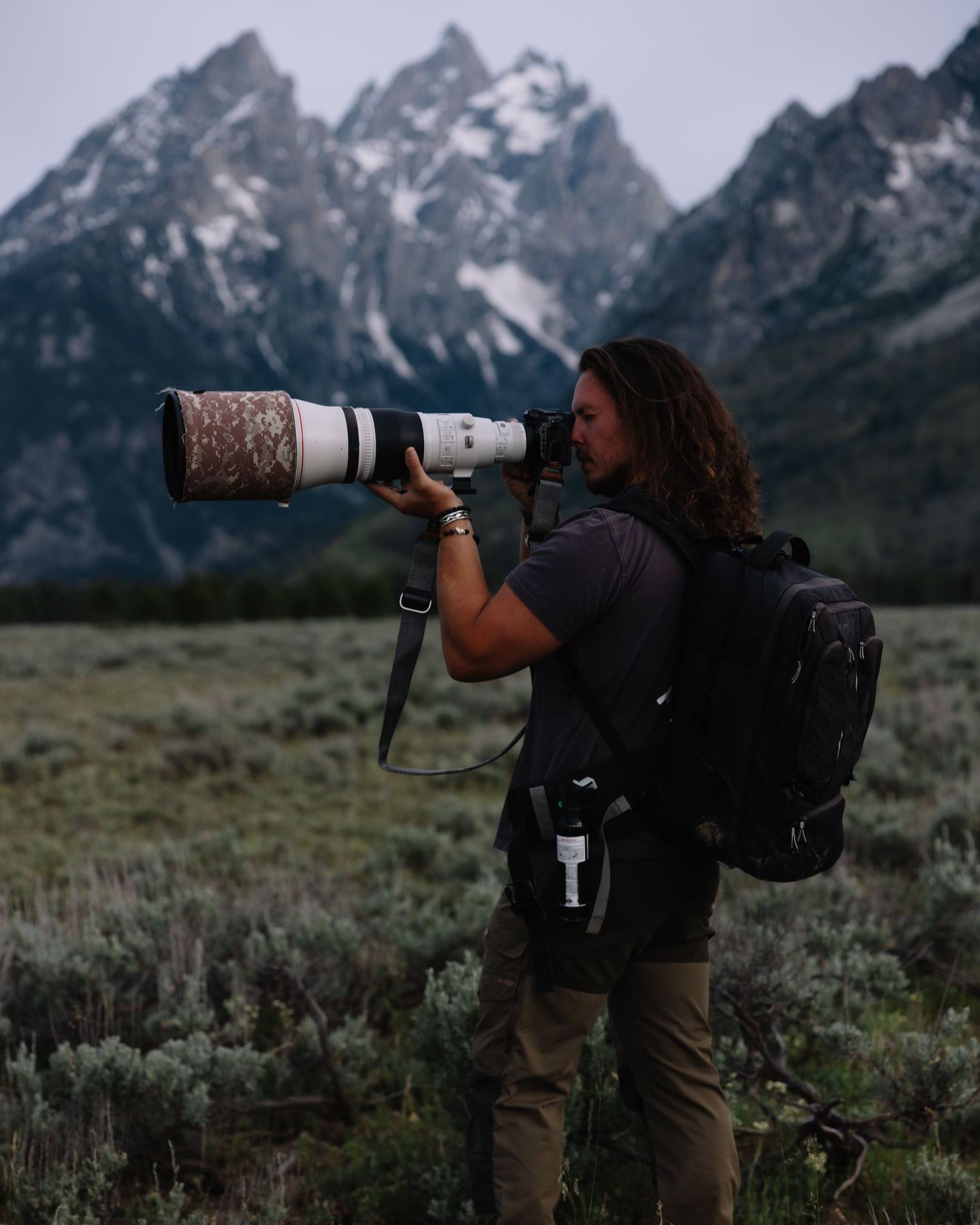 Male photographer with long hair uses large telephoto lens before dramatic snow-capped mountain range.