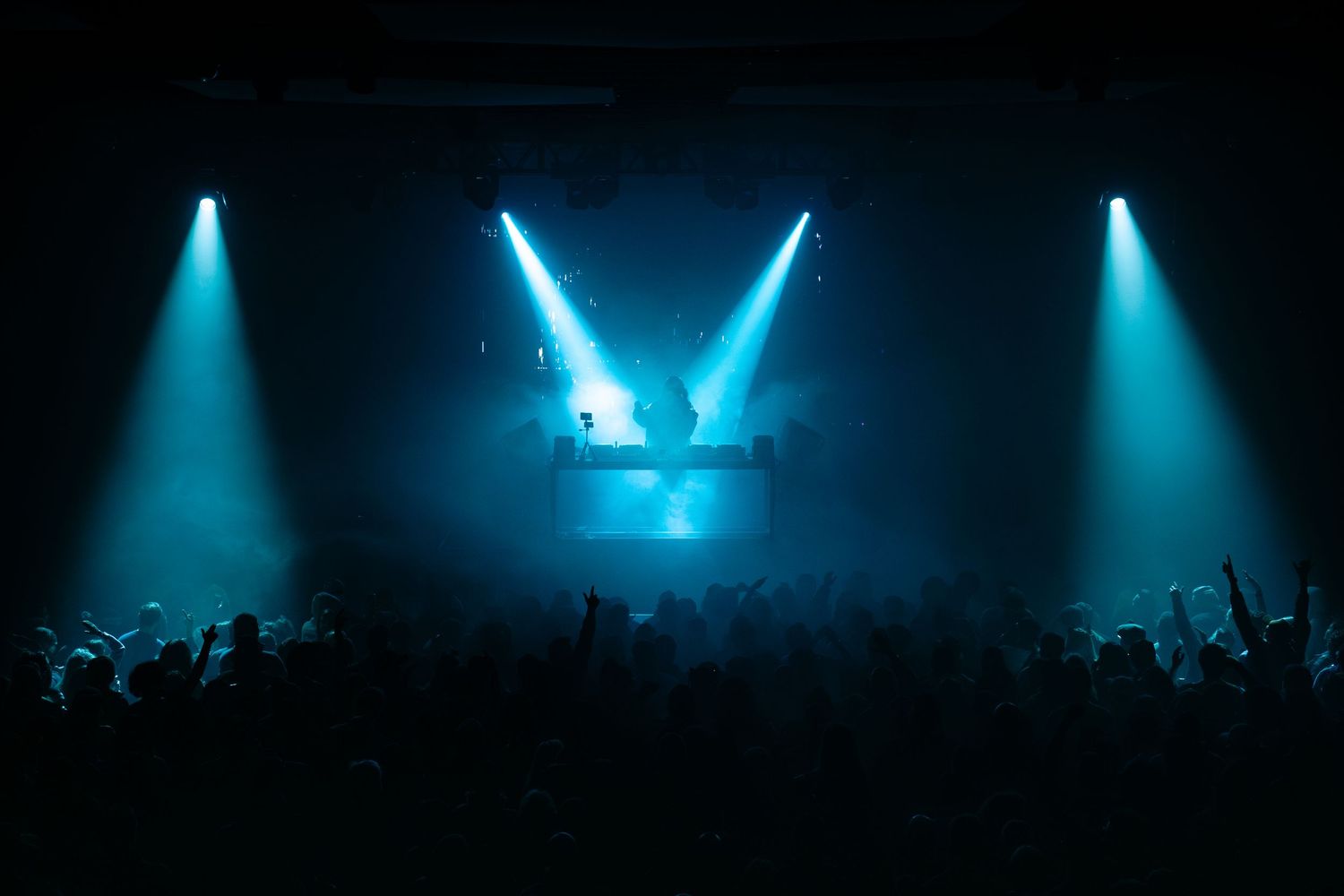 Blue spotlights illuminate a dark concert stage with silhouetted crowd visible in the foreground.