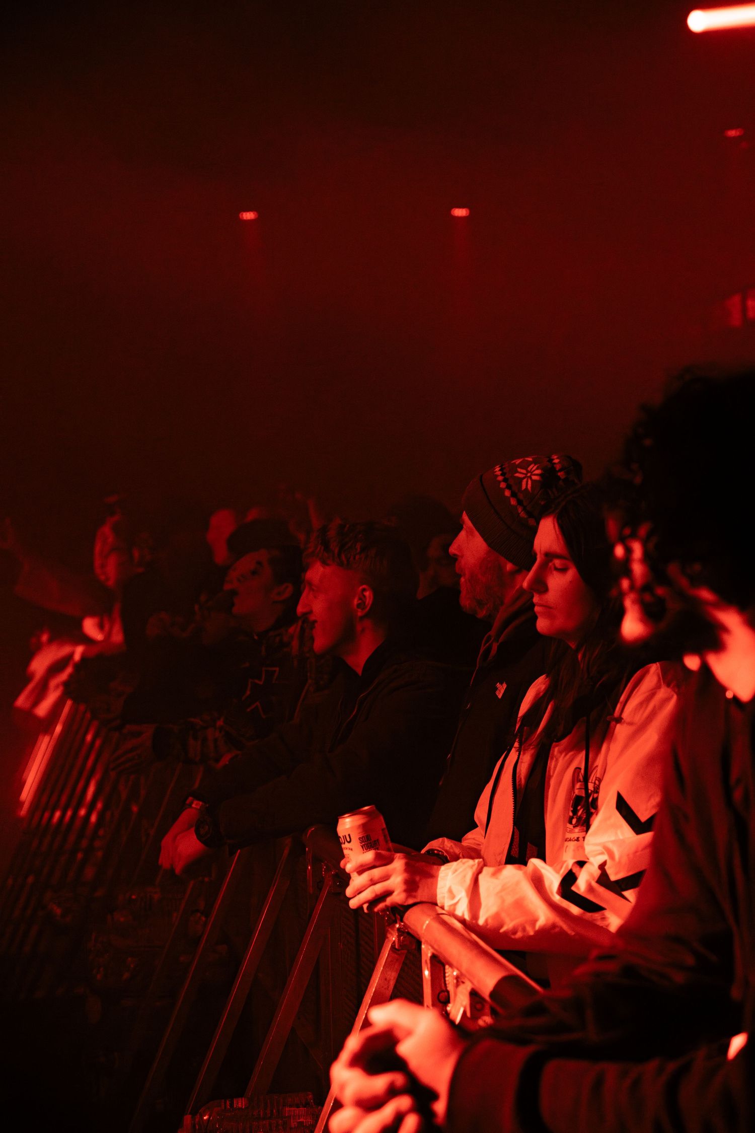 Concert audience silhouettes illuminated by red stage lighting stand watching a performance in a dark venue.