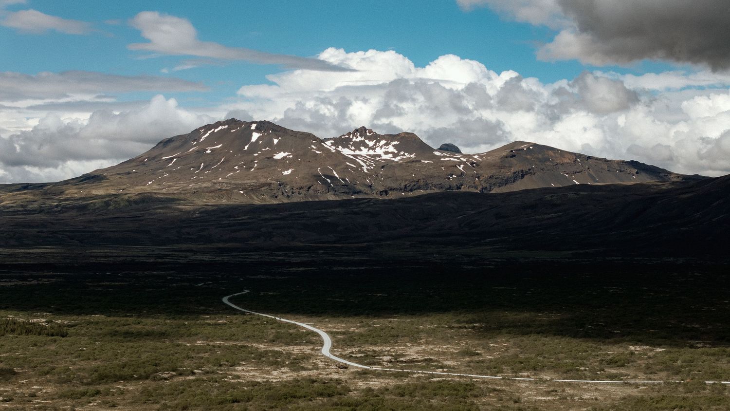 Panoramic mountain landscape with winding road through barren terrain under dramatic clouds.