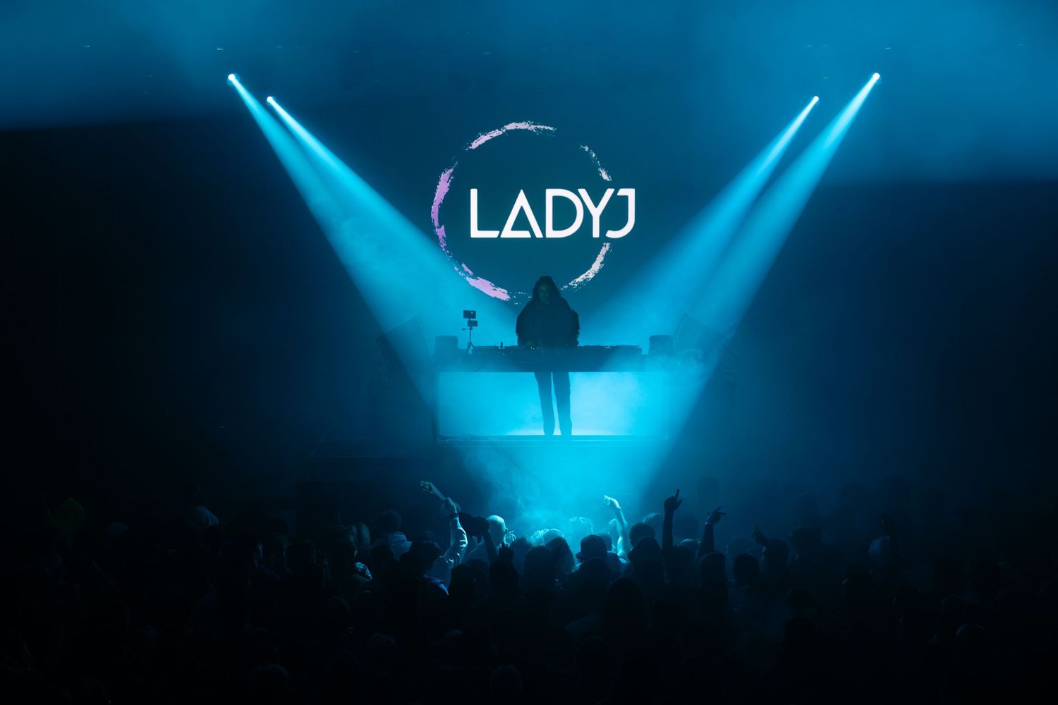 Silhouetted DJ performs on stage with glowing blue LADY sign and spotlights above an energetic crowd.