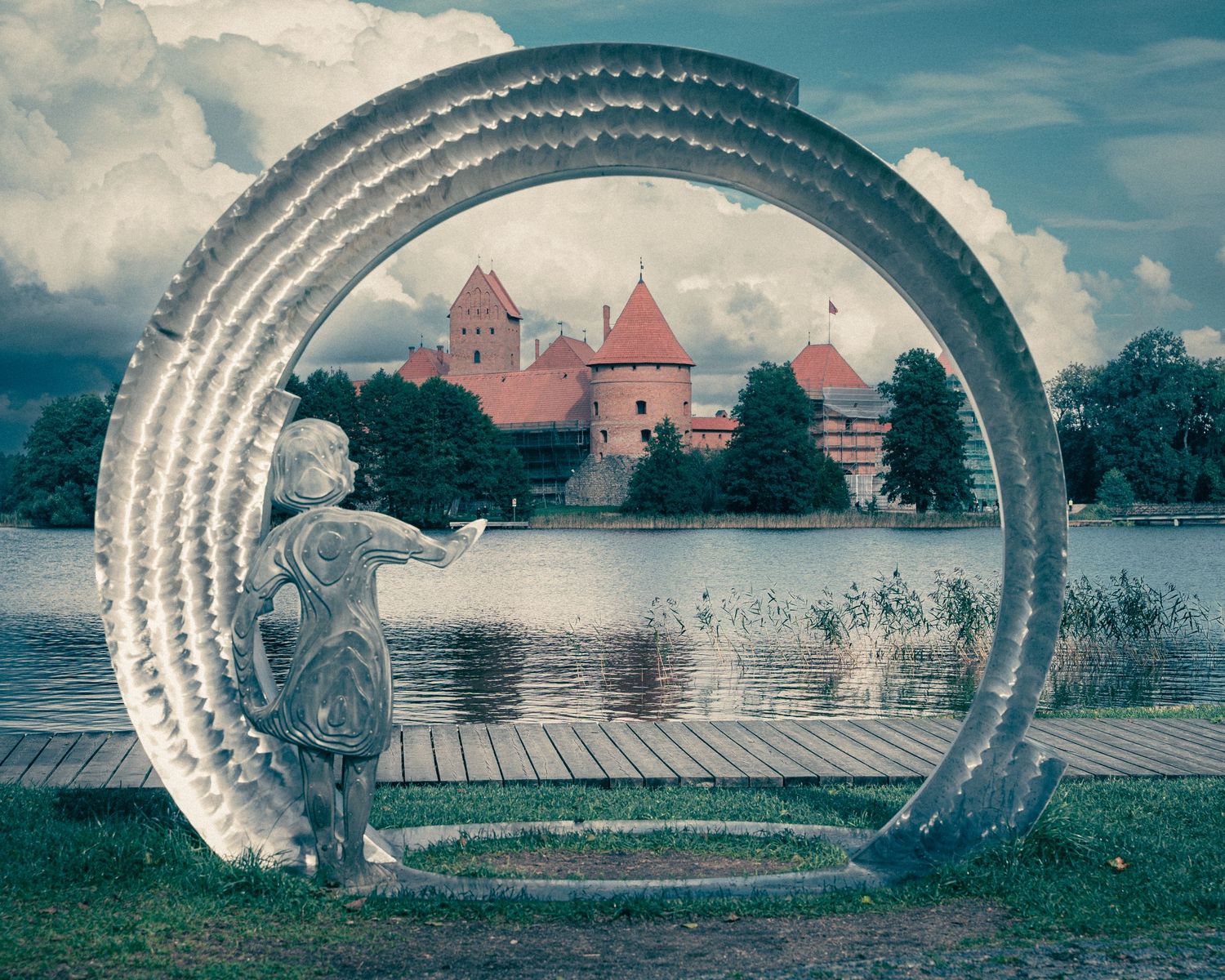 Panoramic view through curved stone archway frames distant castle buildings across a lake.