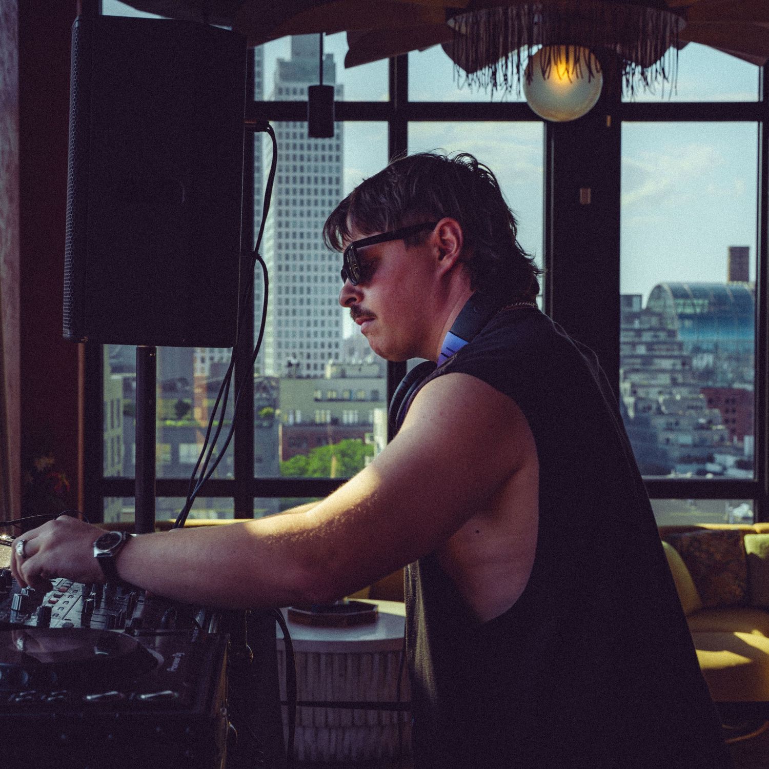 DJ in sleeveless shirt working turntables with city skyline visible through windows.