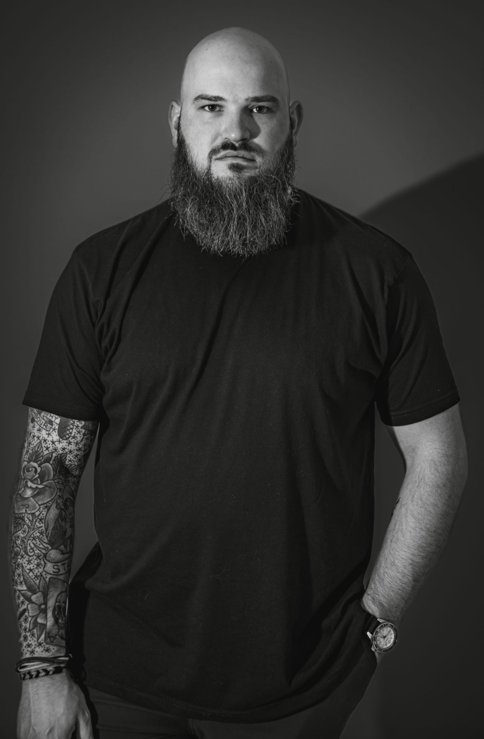 Dramatic monochrome portrait featuring tattooed arm and full beard in studio setting.