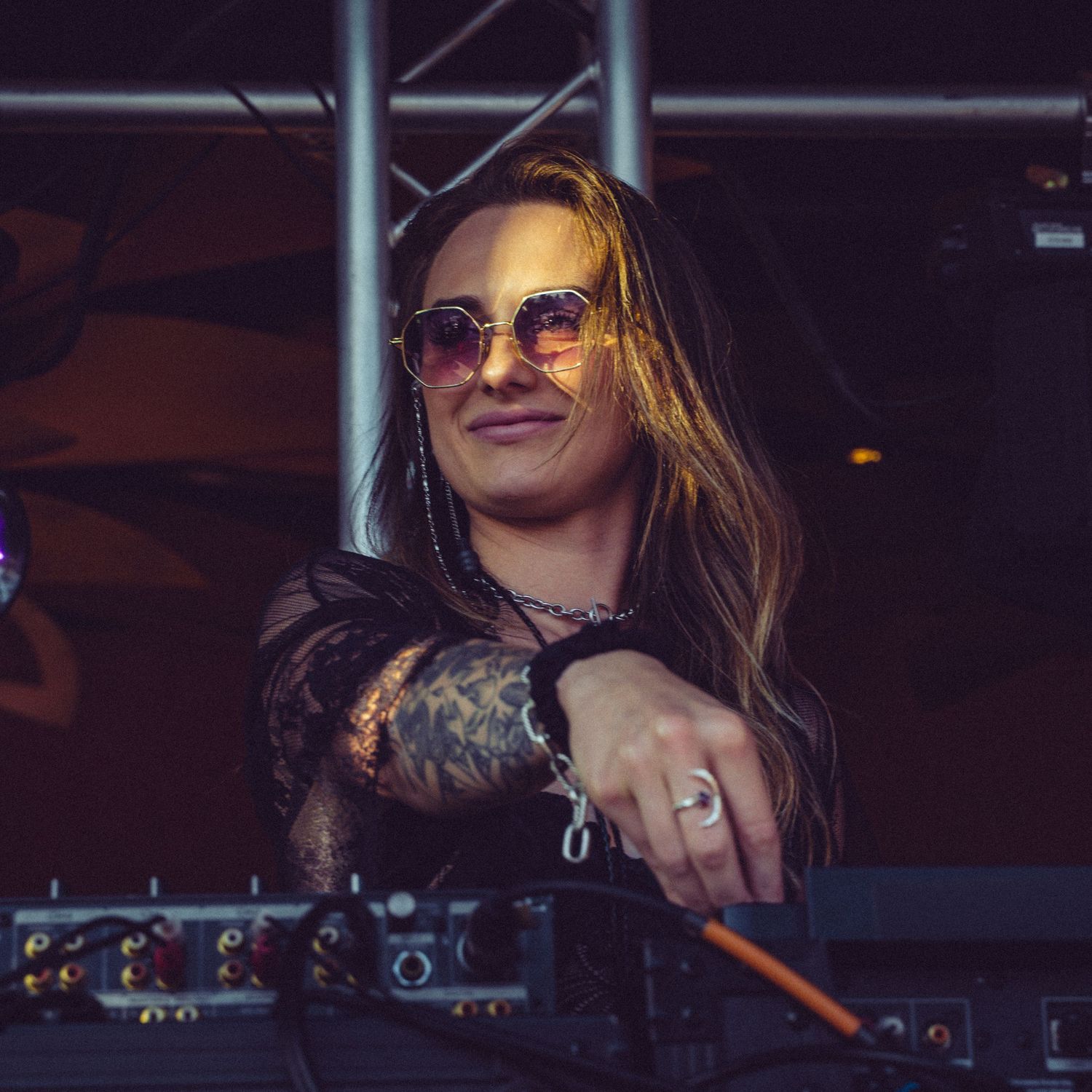 DJ smiling while performing behind mixing equipment at a dark venue with stage lighting.