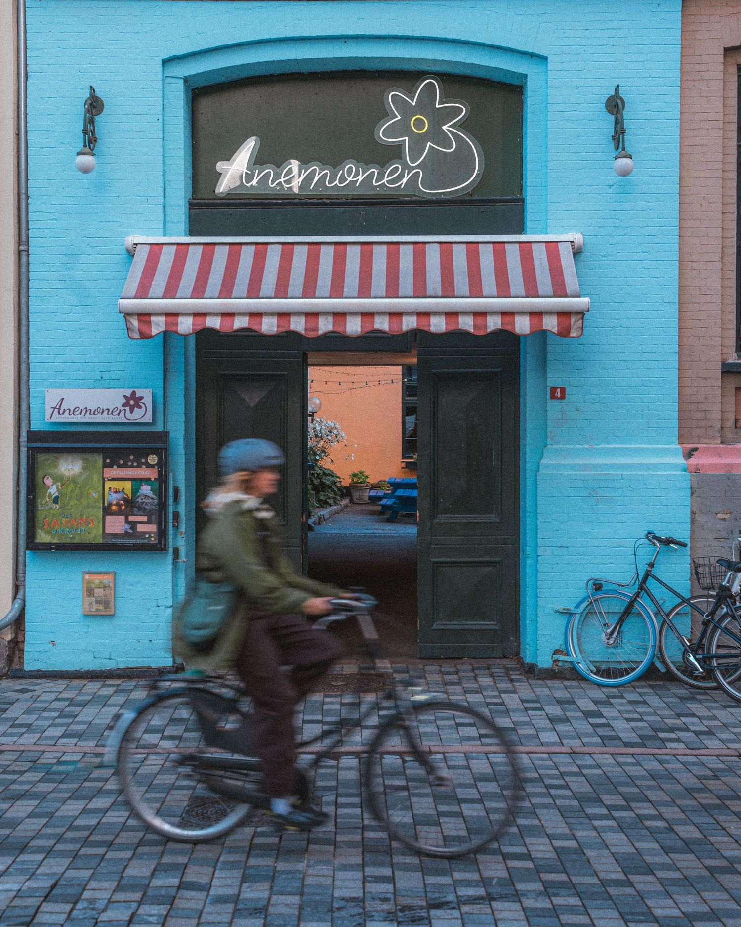 Time-lapse sequence of cyclists passing by a bright blue storefront with neon signage.