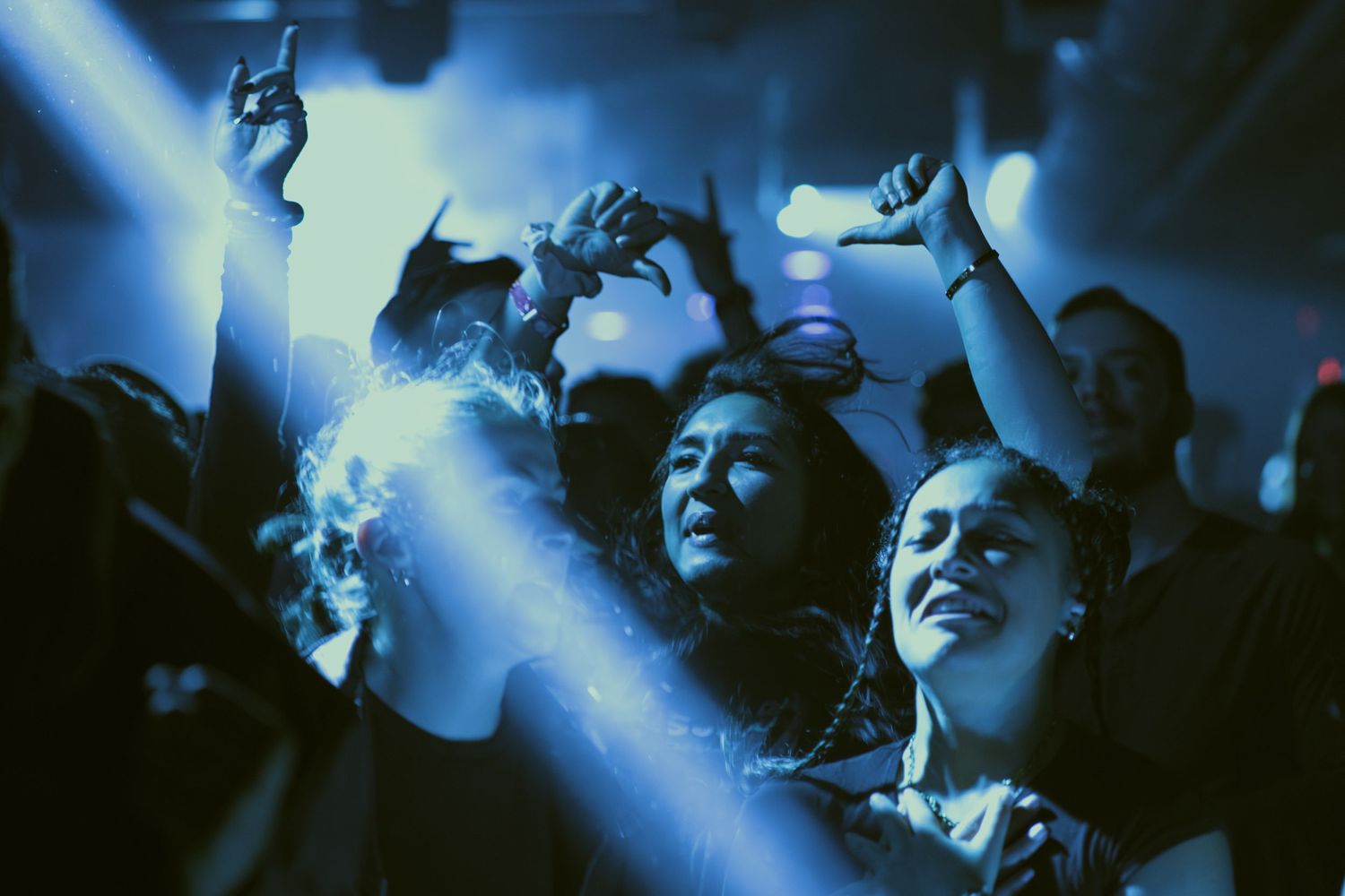 Concert crowd silhouettes illuminated by bright blue spotlights dance with hands raised during a nighttime music performance.
