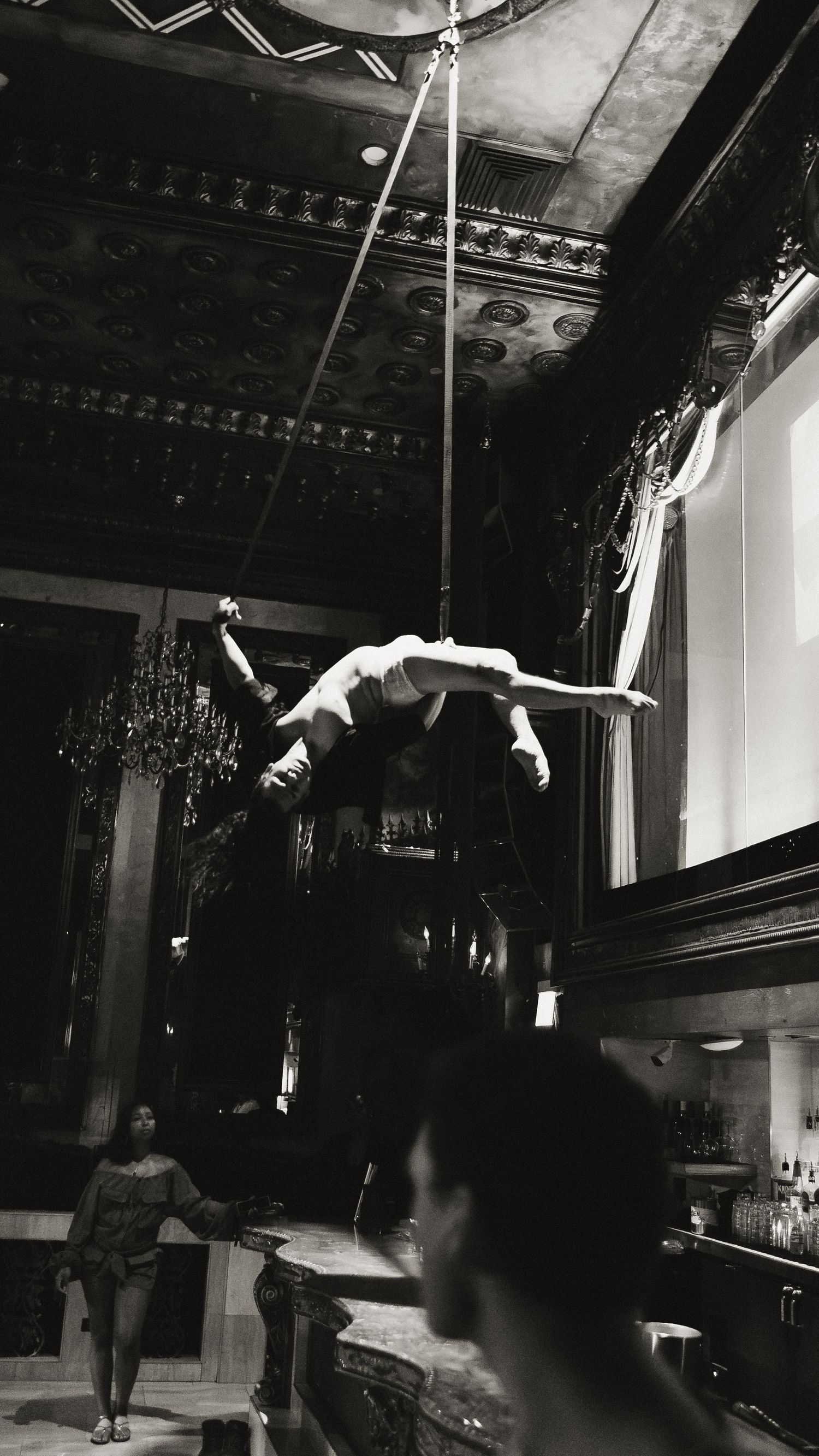 Aerial performers in dark theatrical space practicing acrobatic moves on suspended equipment.