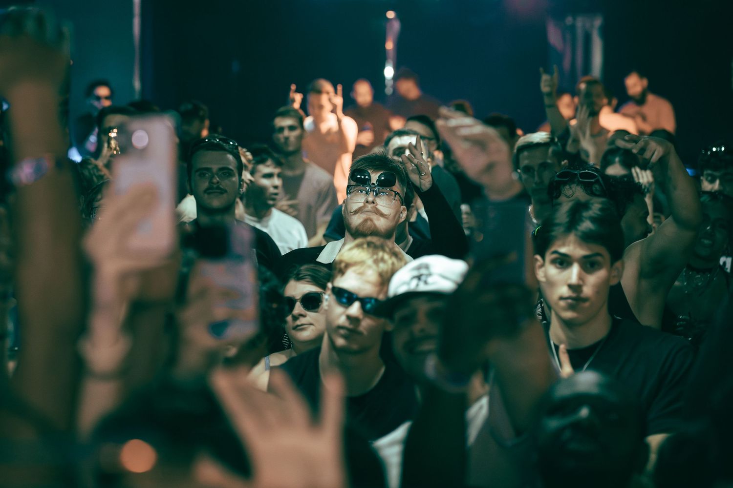 A crowded concert audience in dark lighting with phones raised and people packed together in the crowd.