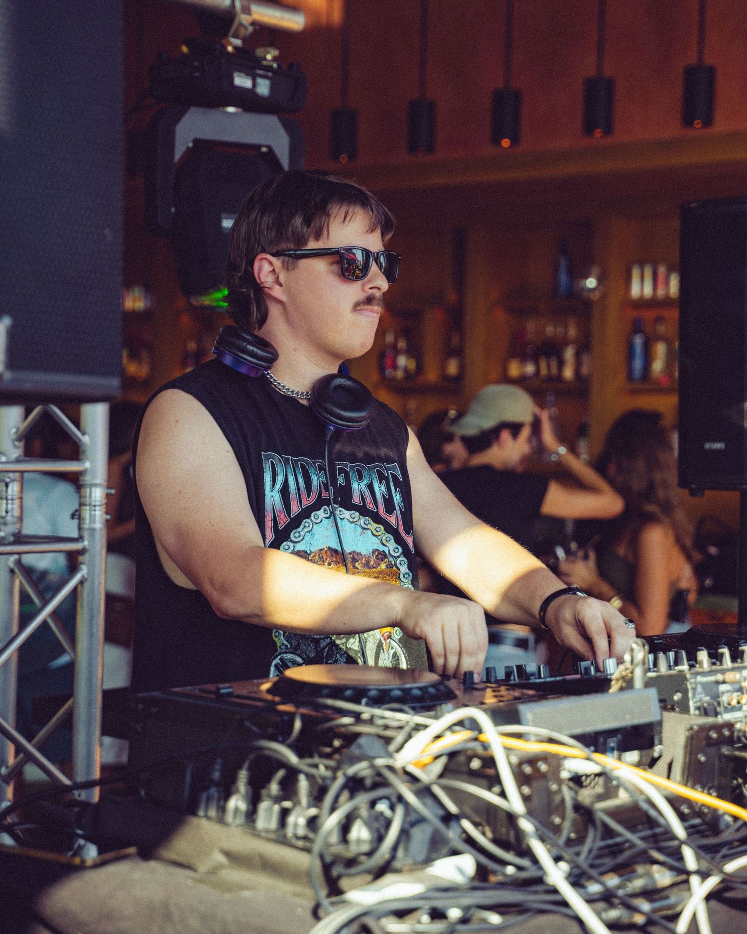 DJ wearing sunglasses performs at mixing board in dimly lit club setting.