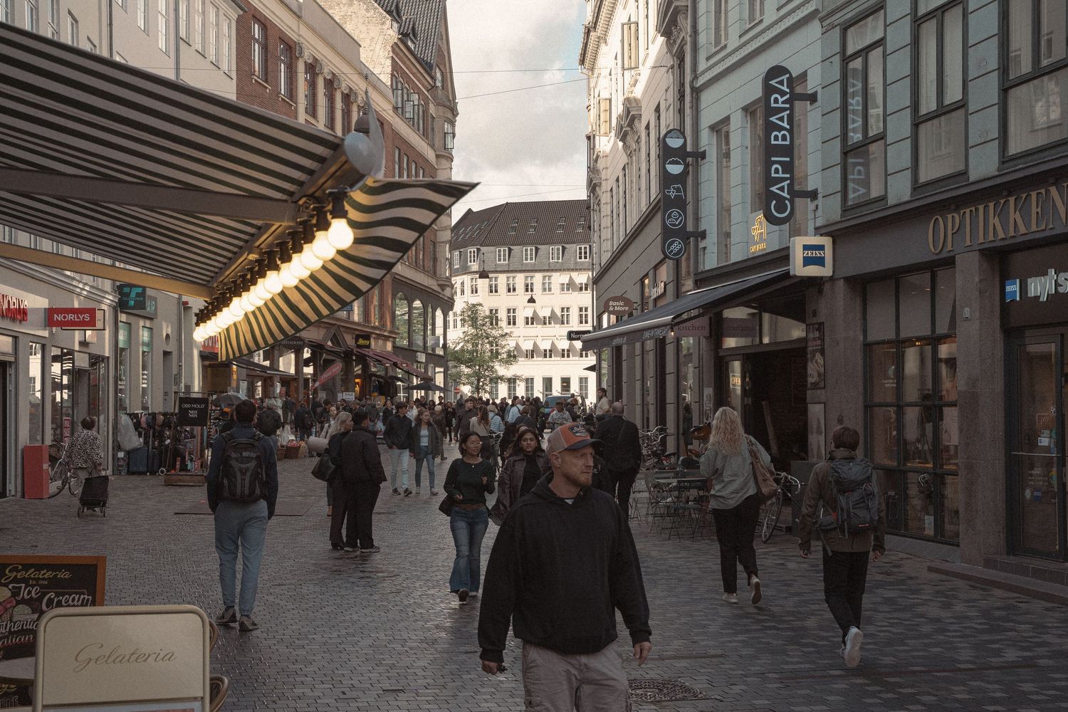 Busy European city street with pedestrians walking along storefronts at dusk.