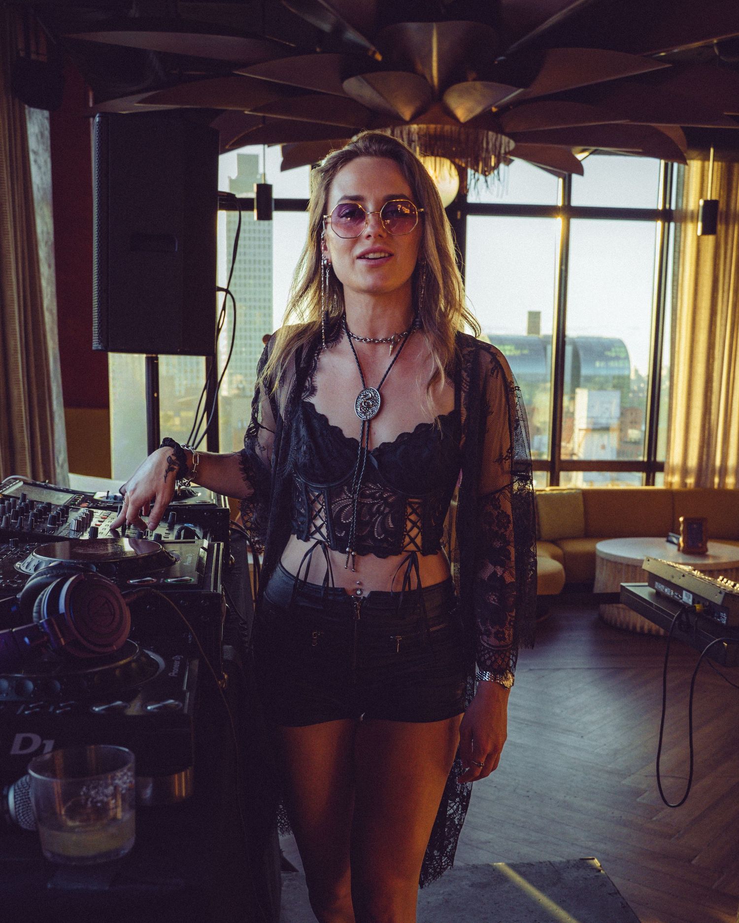 DJ in black lace outfit performing at rooftop venue with city views.