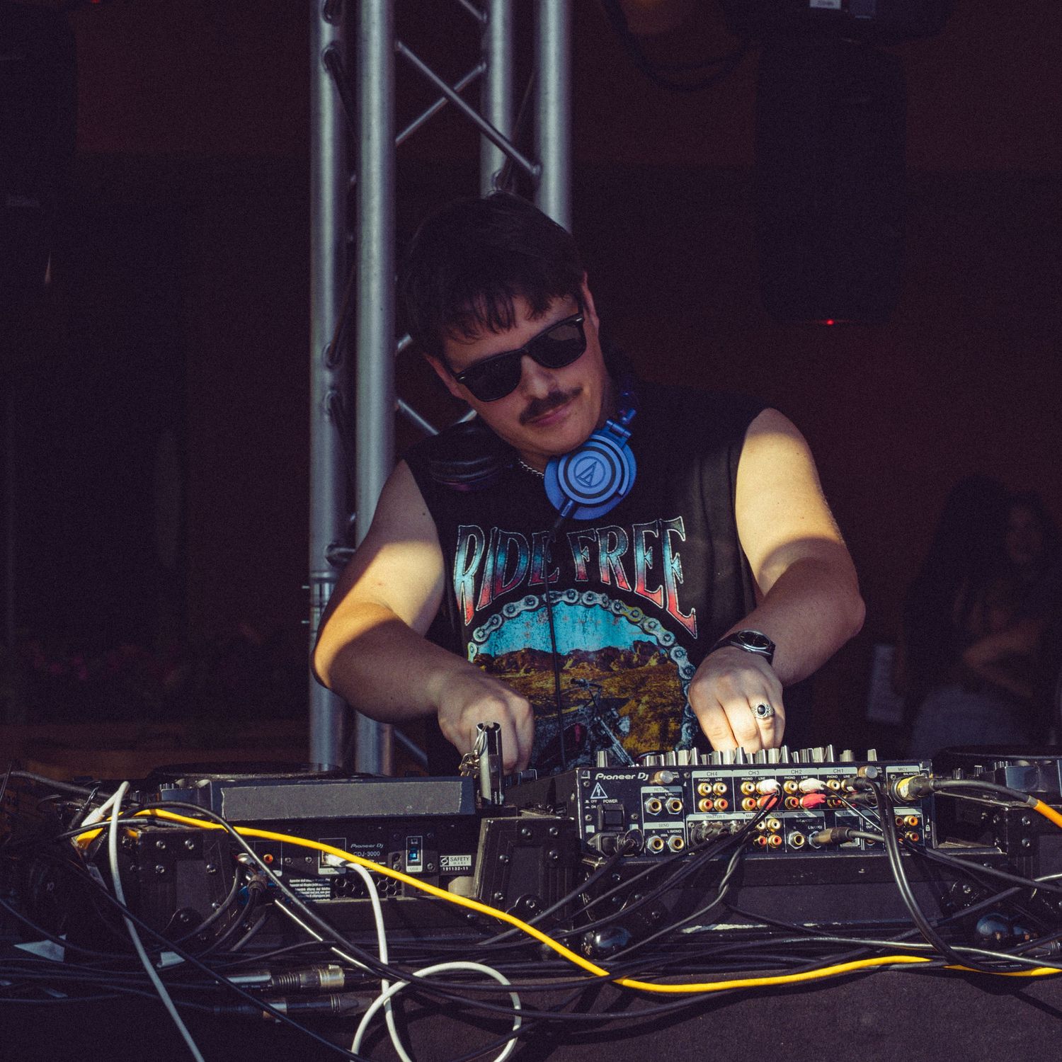 DJ in sleeveless band shirt mixing music at dark indoor venue.