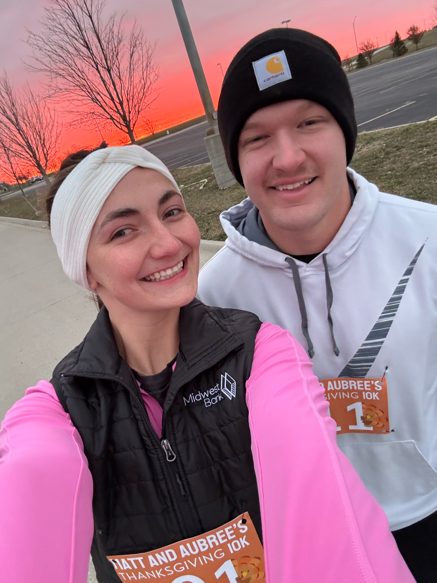Two people wearing winter hats pose for selfies outdoors during a vibrant pink and orange sunset.