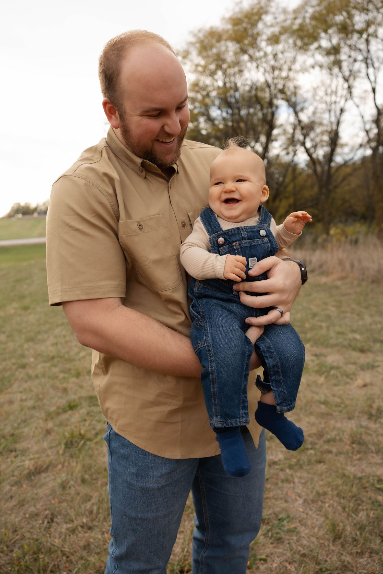 A baby in denim overalls shares a playful moment with an adult in a tan shirt outdoors.