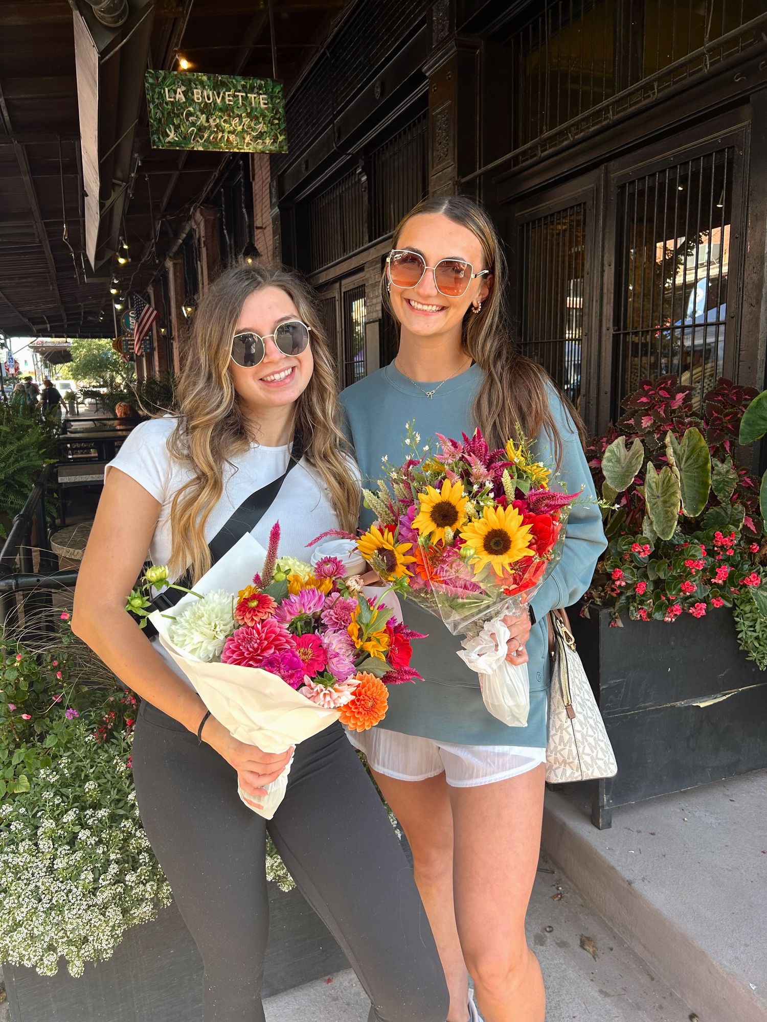 People holding vibrant bouquets of fresh flowers including sunflowers and wildflowers outside a rustic wooden storefront.