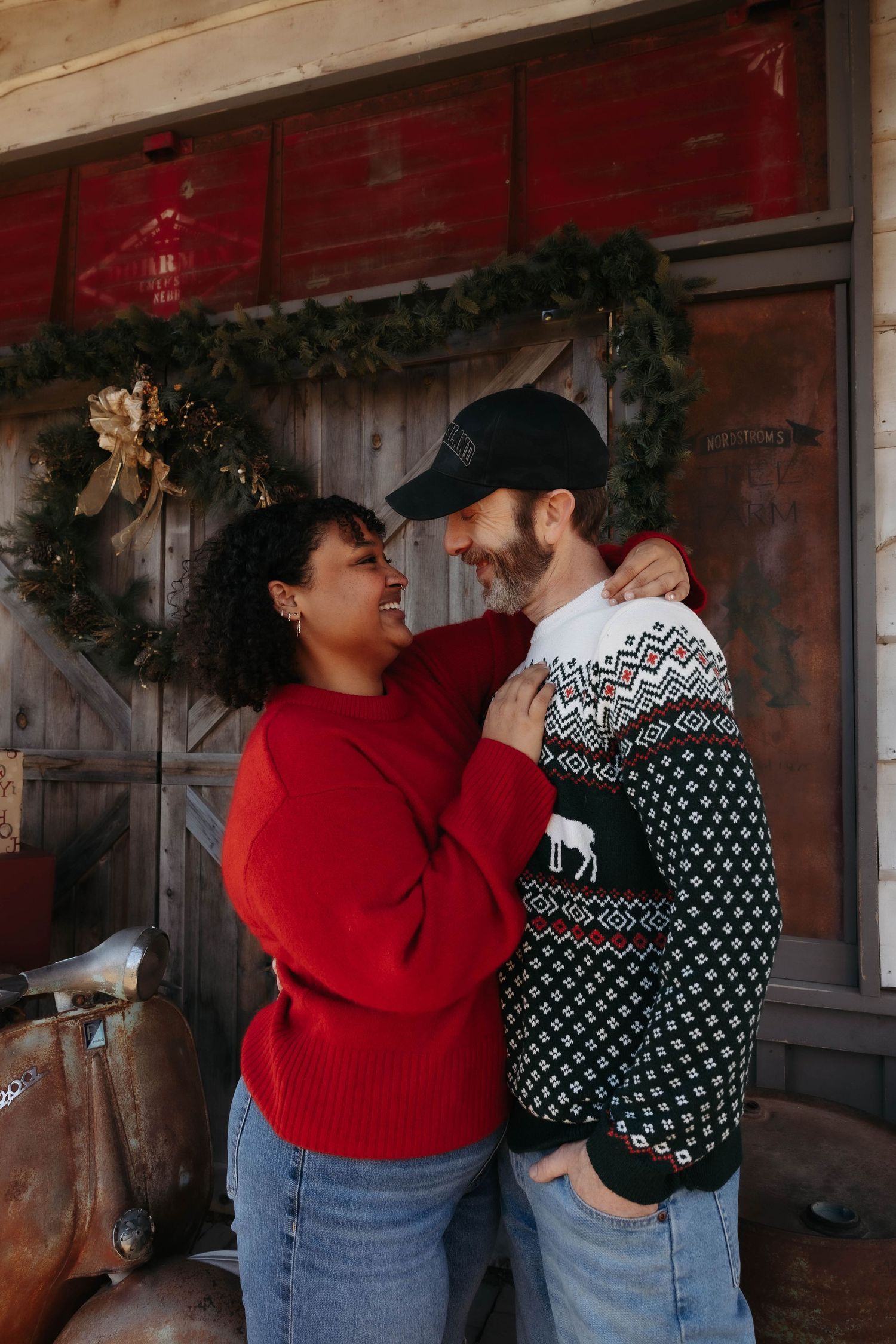 Couple in festive sweaters share a romantic moment by rustic barn door with holiday decorations.