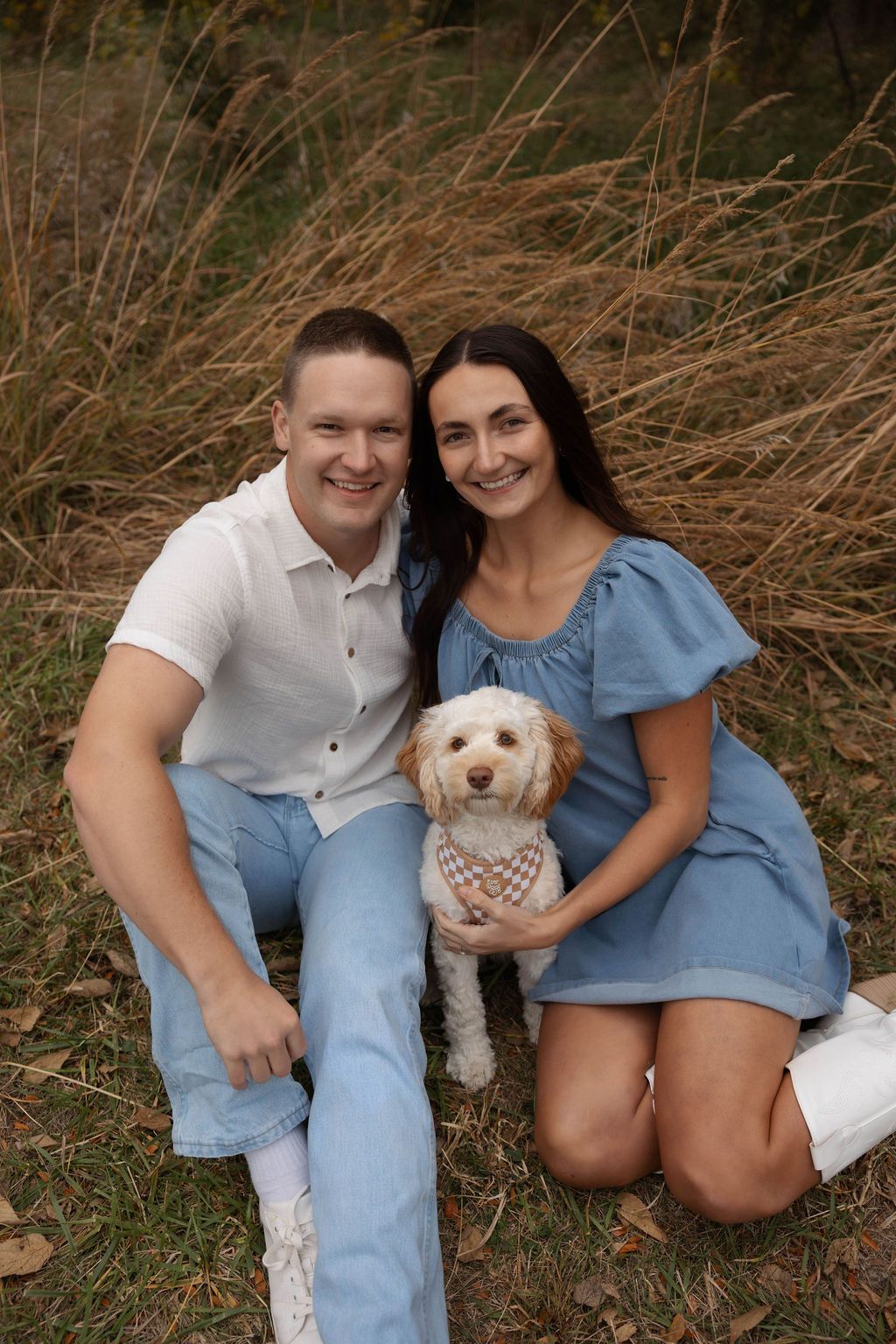 A couple sits in tall grass with their small white goldendoodle puppy wearing a bandana during an outdoor photo session.