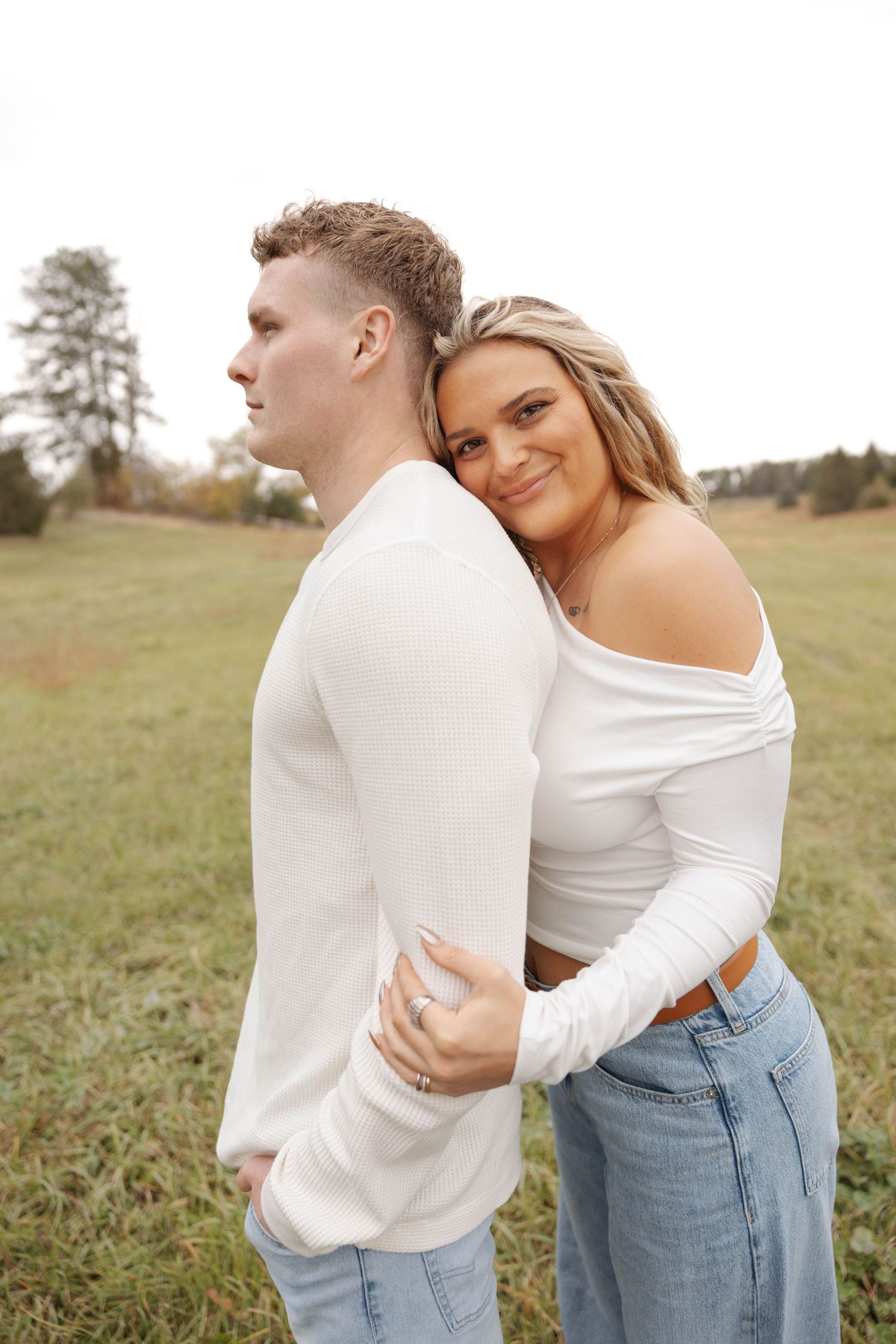 Couple in white tops and jeans embraces in outdoor field setting.