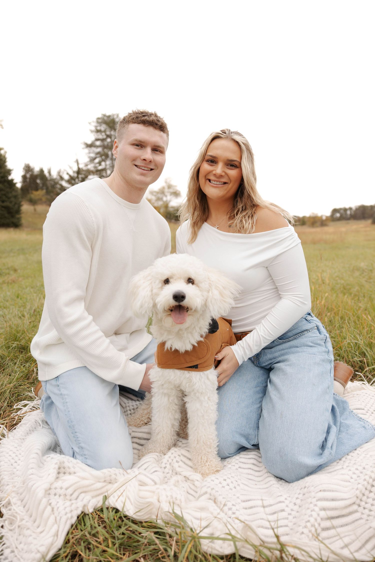 Couple in casual white and denim outfits sits on blanket with white fluffy dog.