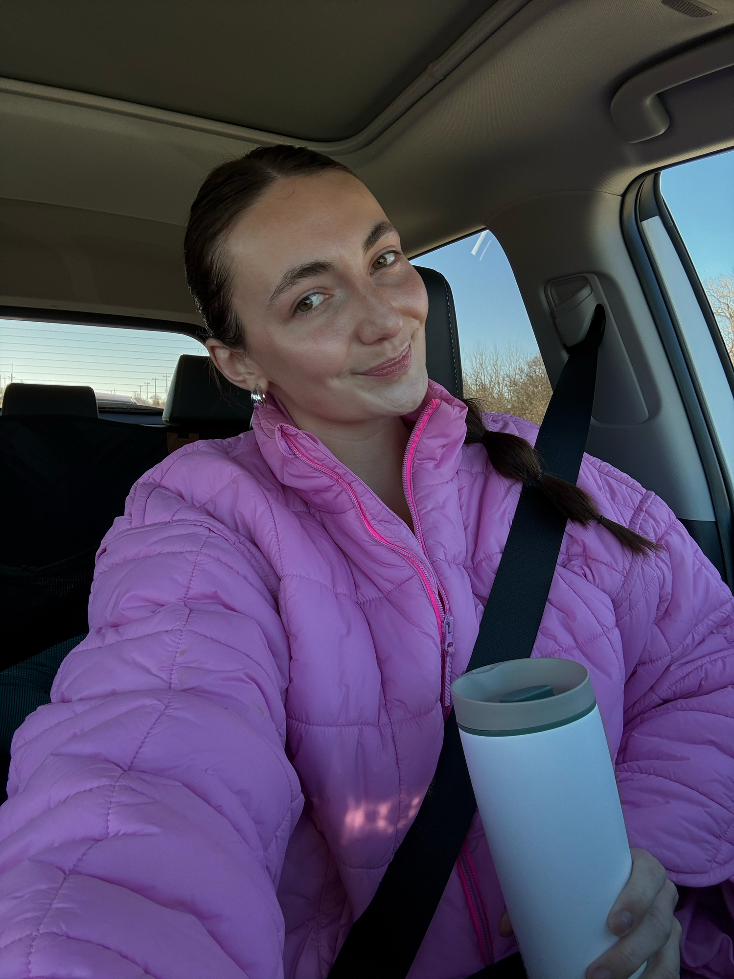 Person in a bright pink puffer jacket takes a selfie while sitting in a car with a reusable drink container.