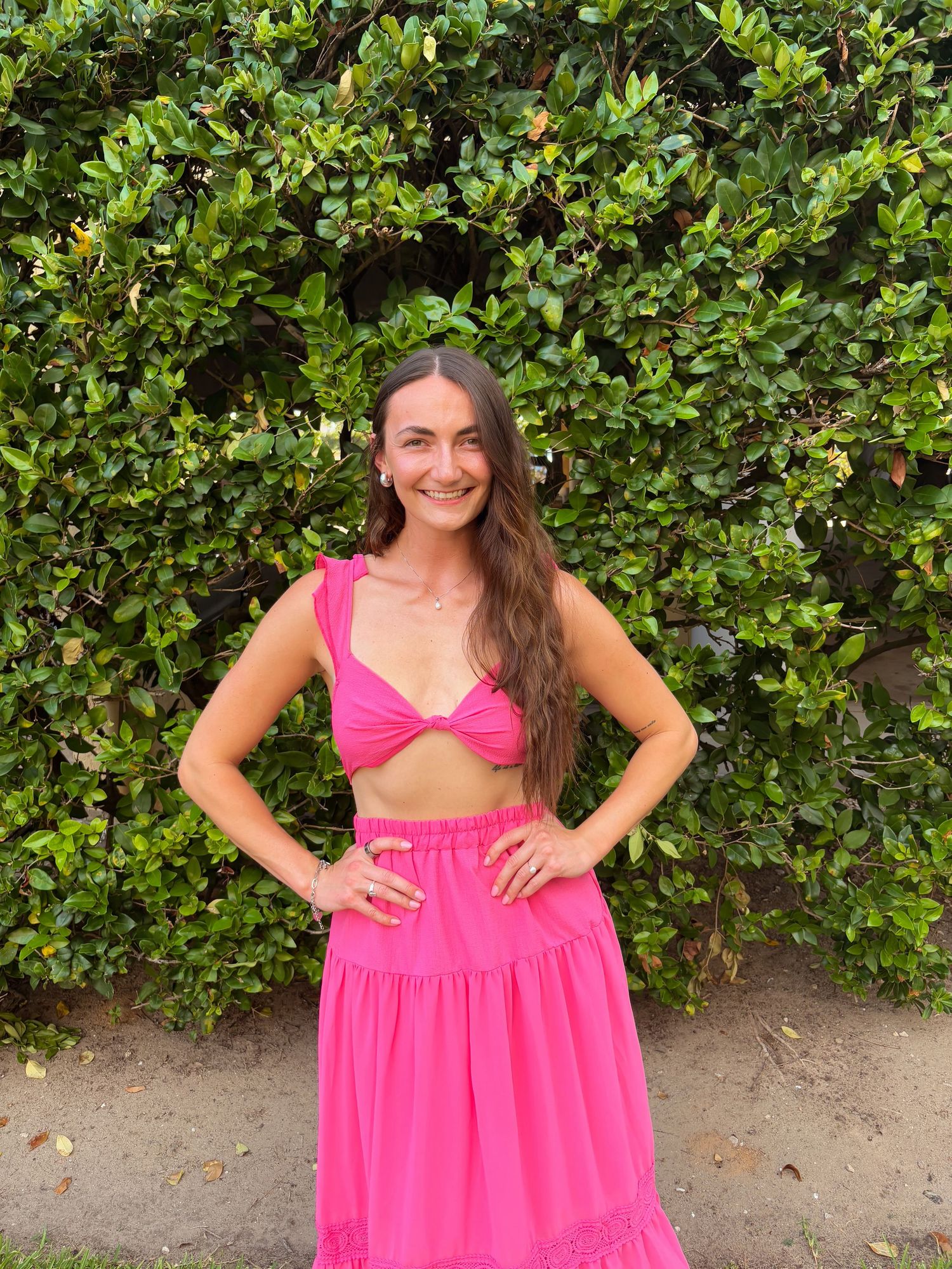 Woman in bright pink summer dress poses against green hedge backdrop.