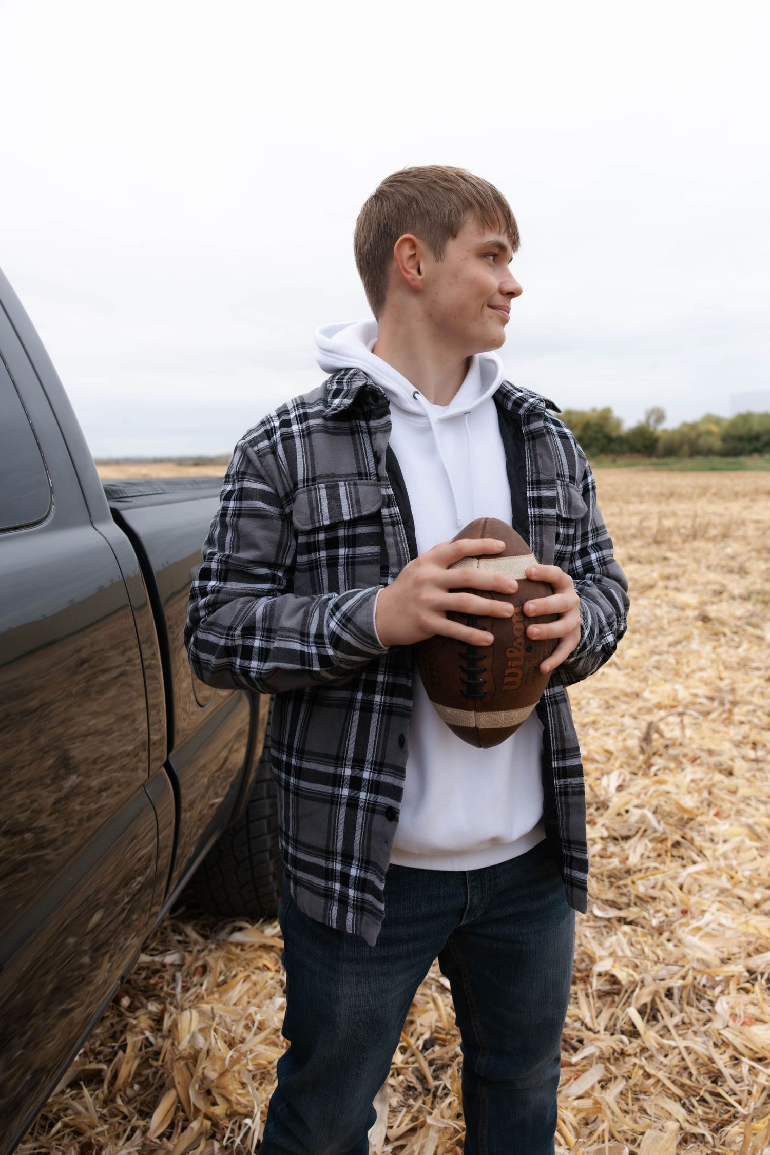 Someone in a plaid shirt and jeans stands in a harvested cornfield holding a football.