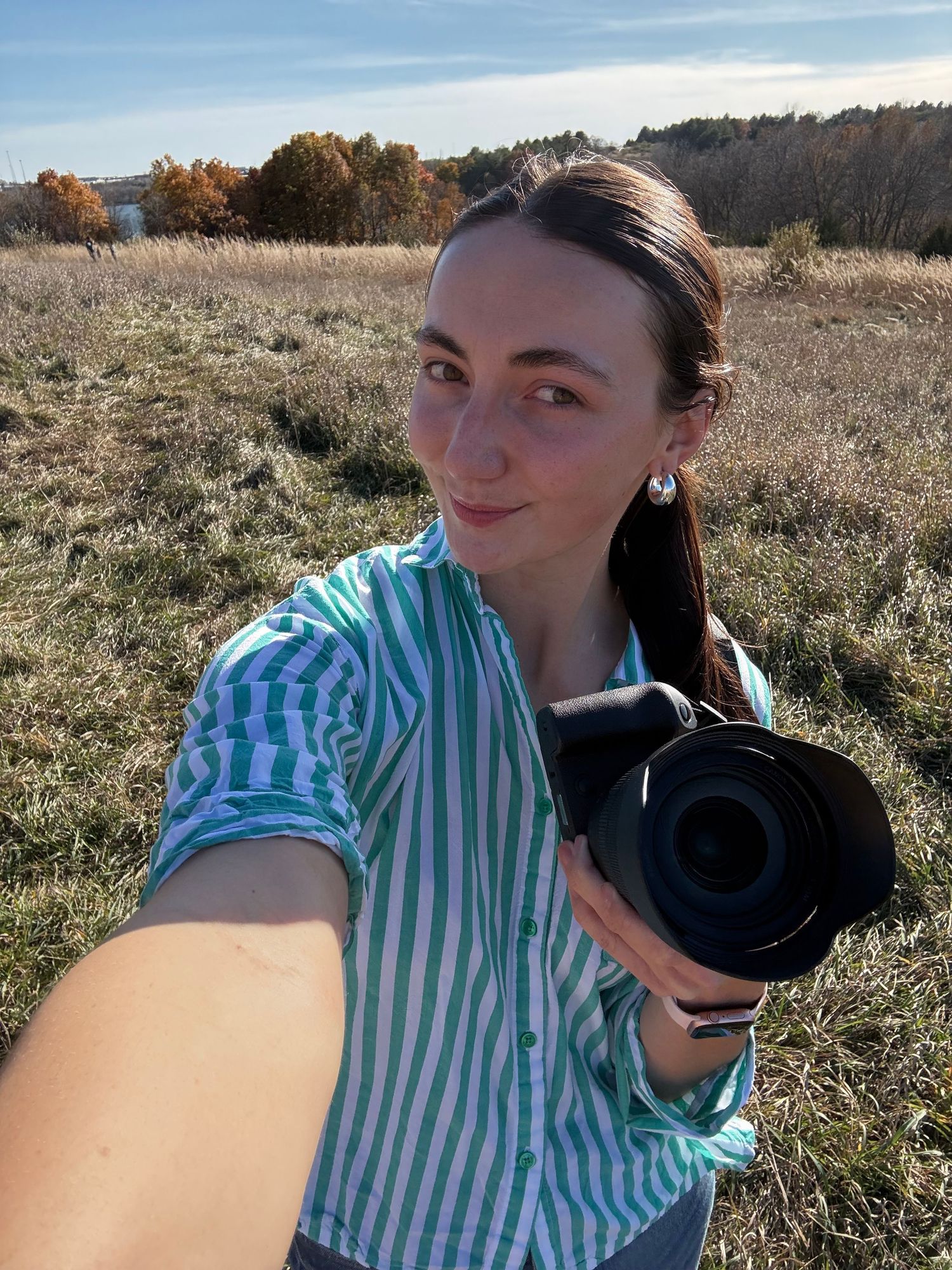 Woman with camera poses in blue striped shirt against autumn landscape.