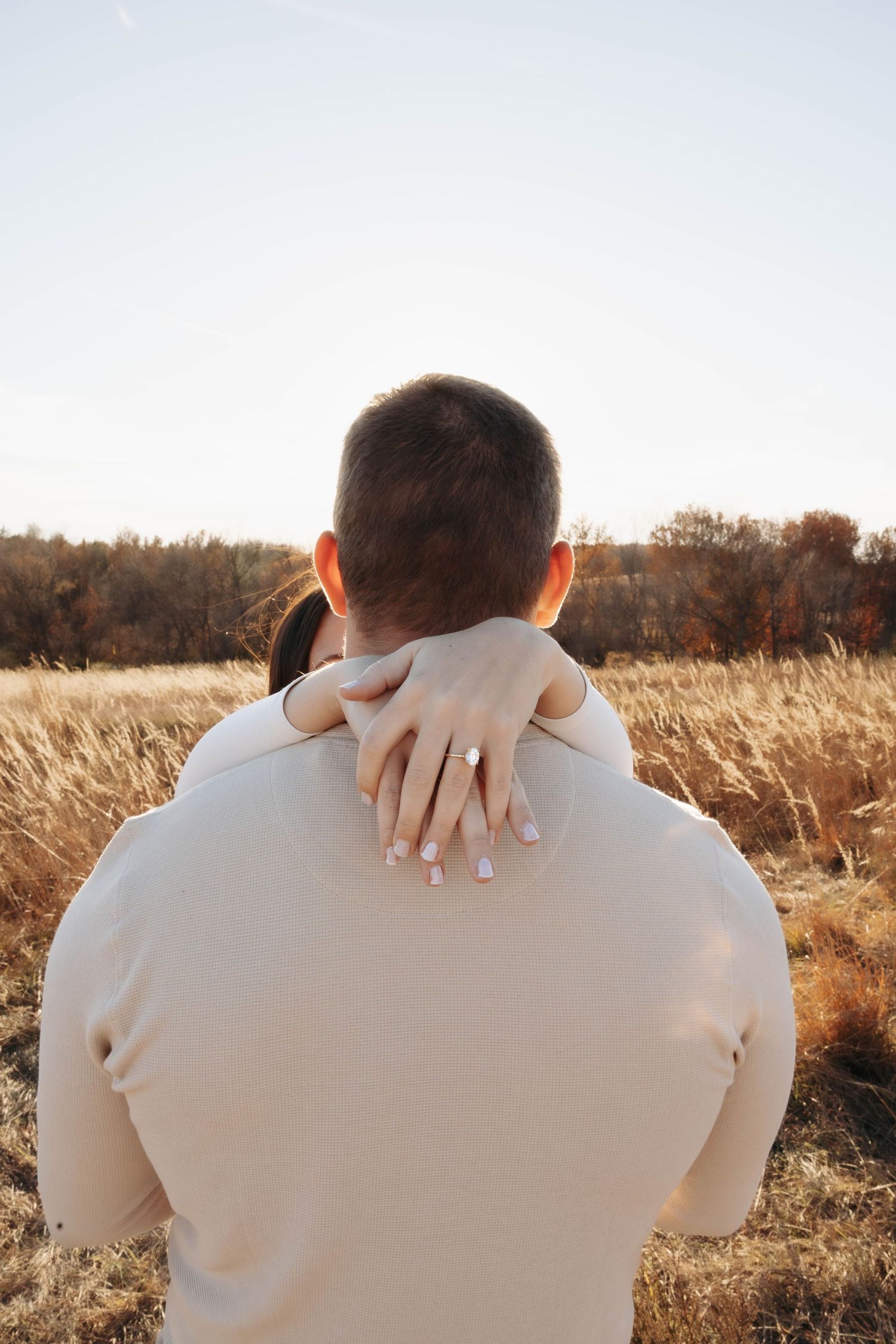 Back view of embrace with hands showing engagement ring in golden field.