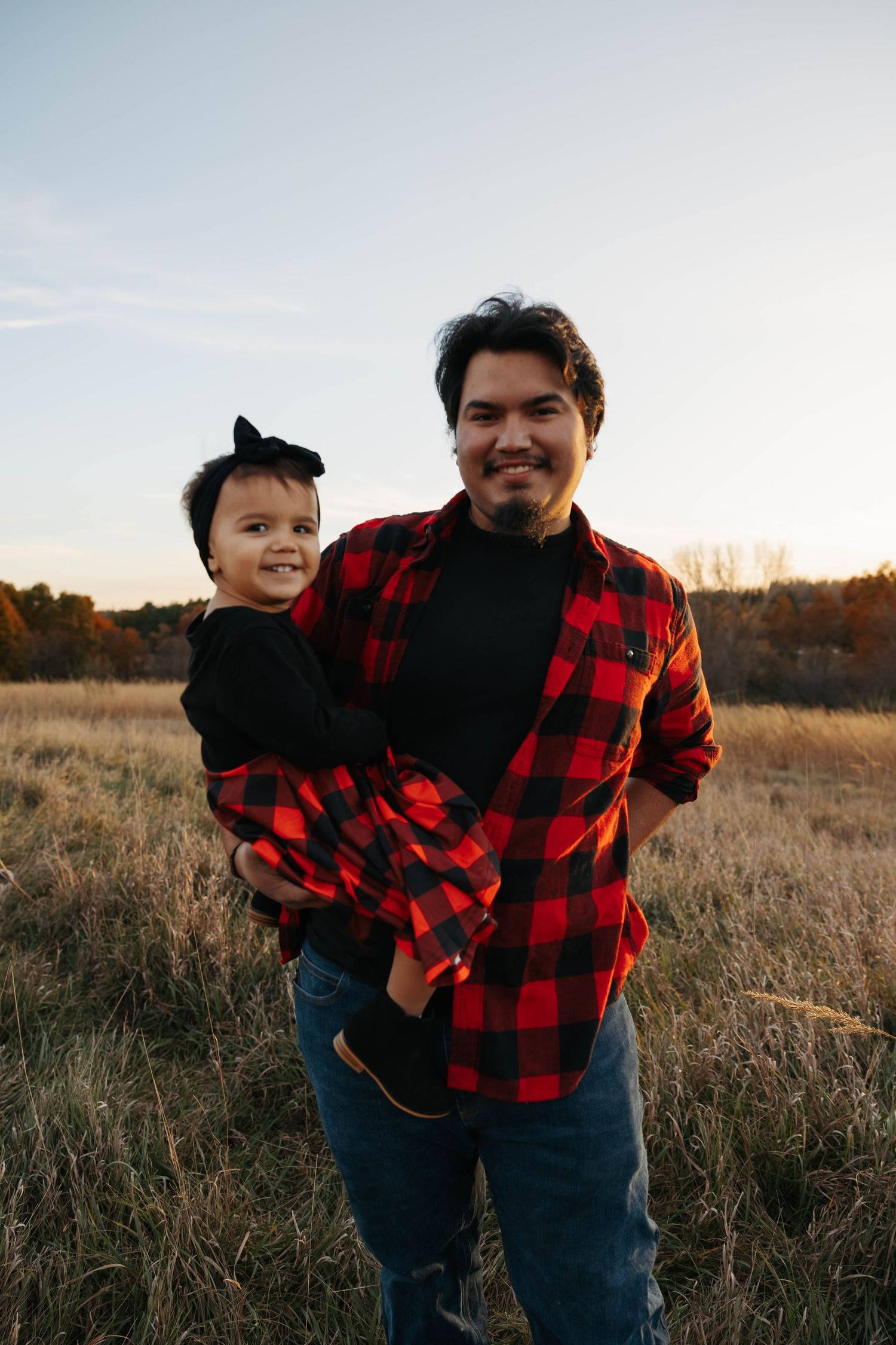 A family wearing matching buffalo plaid shirts poses together in a sunset-lit field.