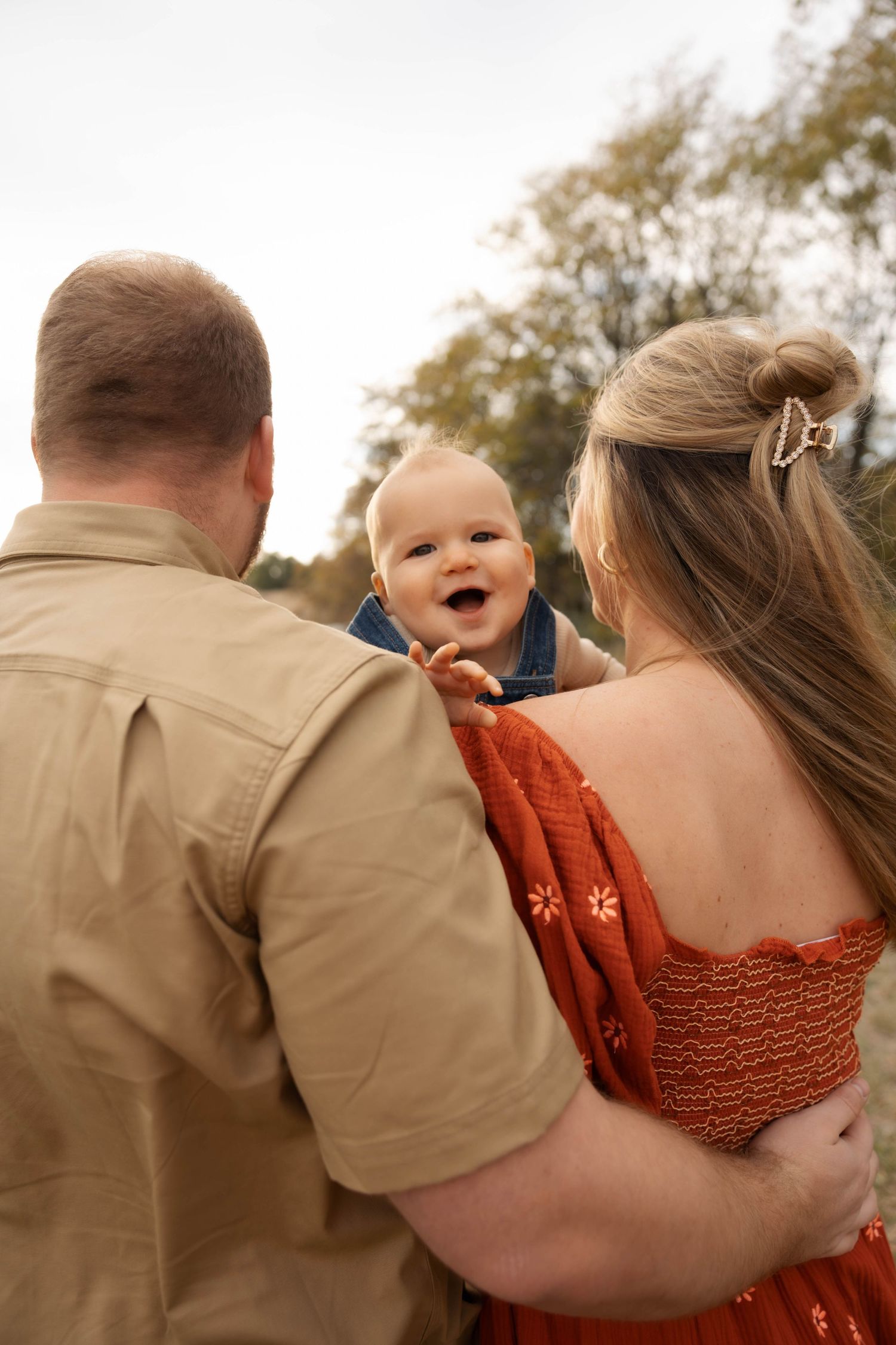 A couple in casual attire embraces while holding their baby during an outdoor photo session.