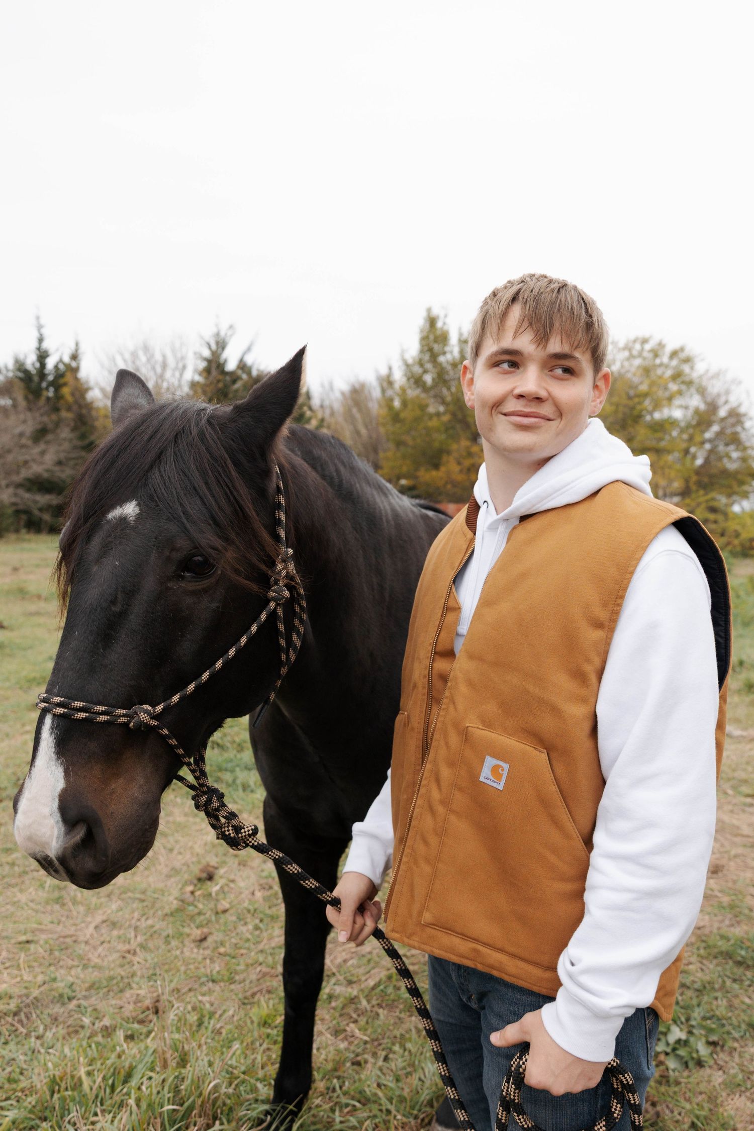 A black horse with braided mane stands next to someone wearing a brown vest and white shirt in an autumn field.