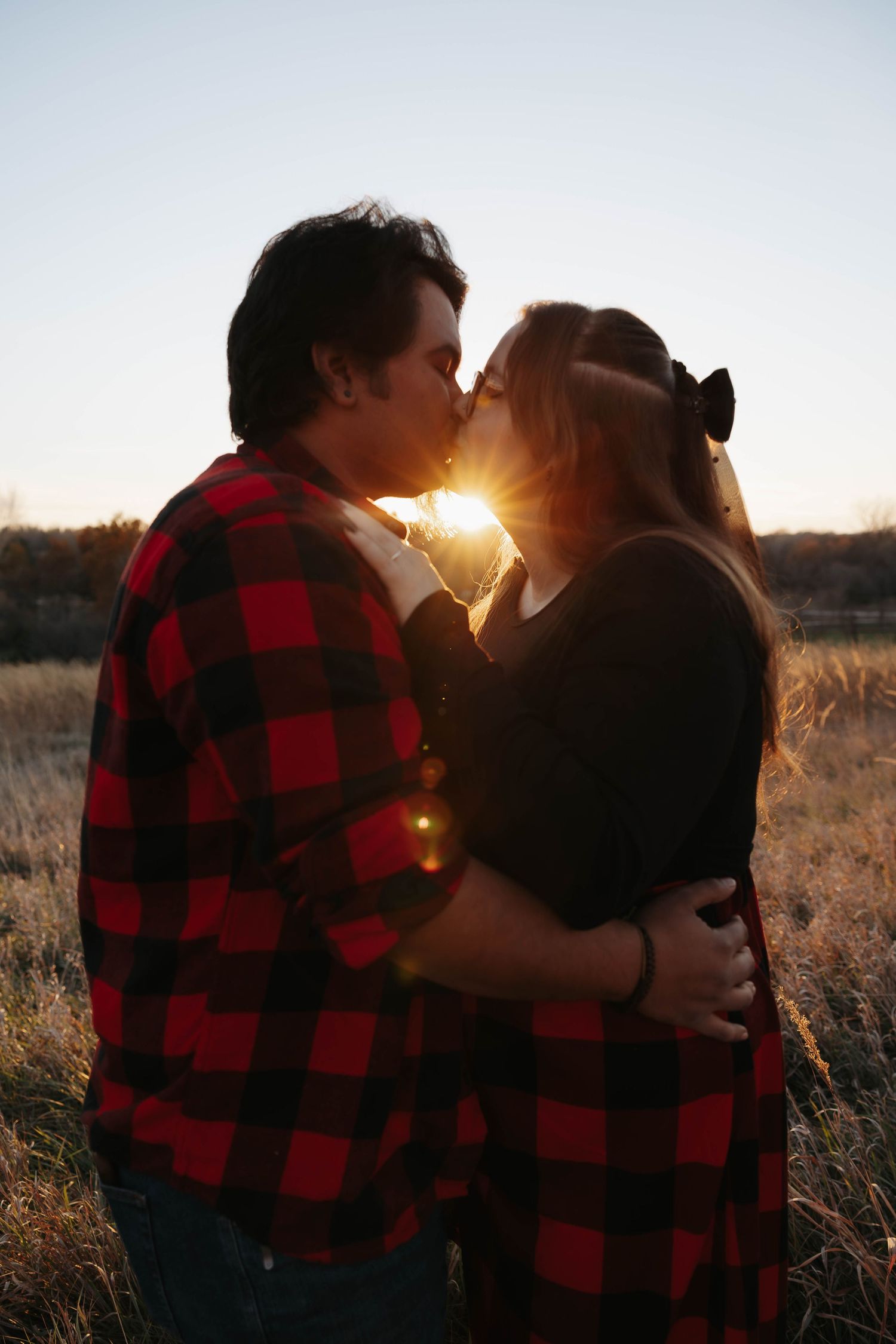 A couple in buffalo plaid and black clothing embraces in a field at sunset.