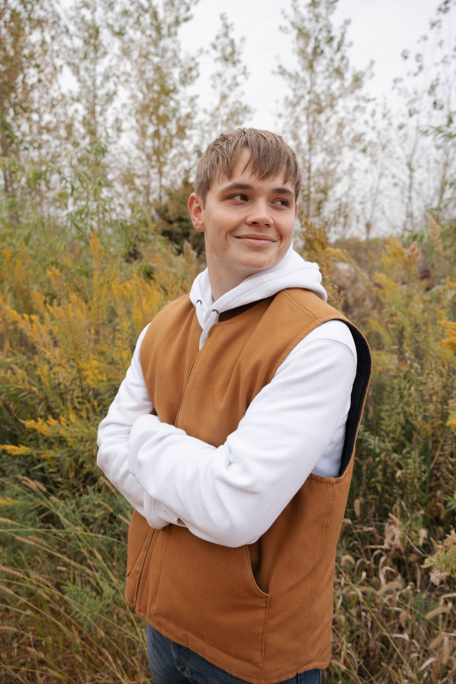 Someone wearing a brown vest and white sweater poses outdoors against autumn foliage.